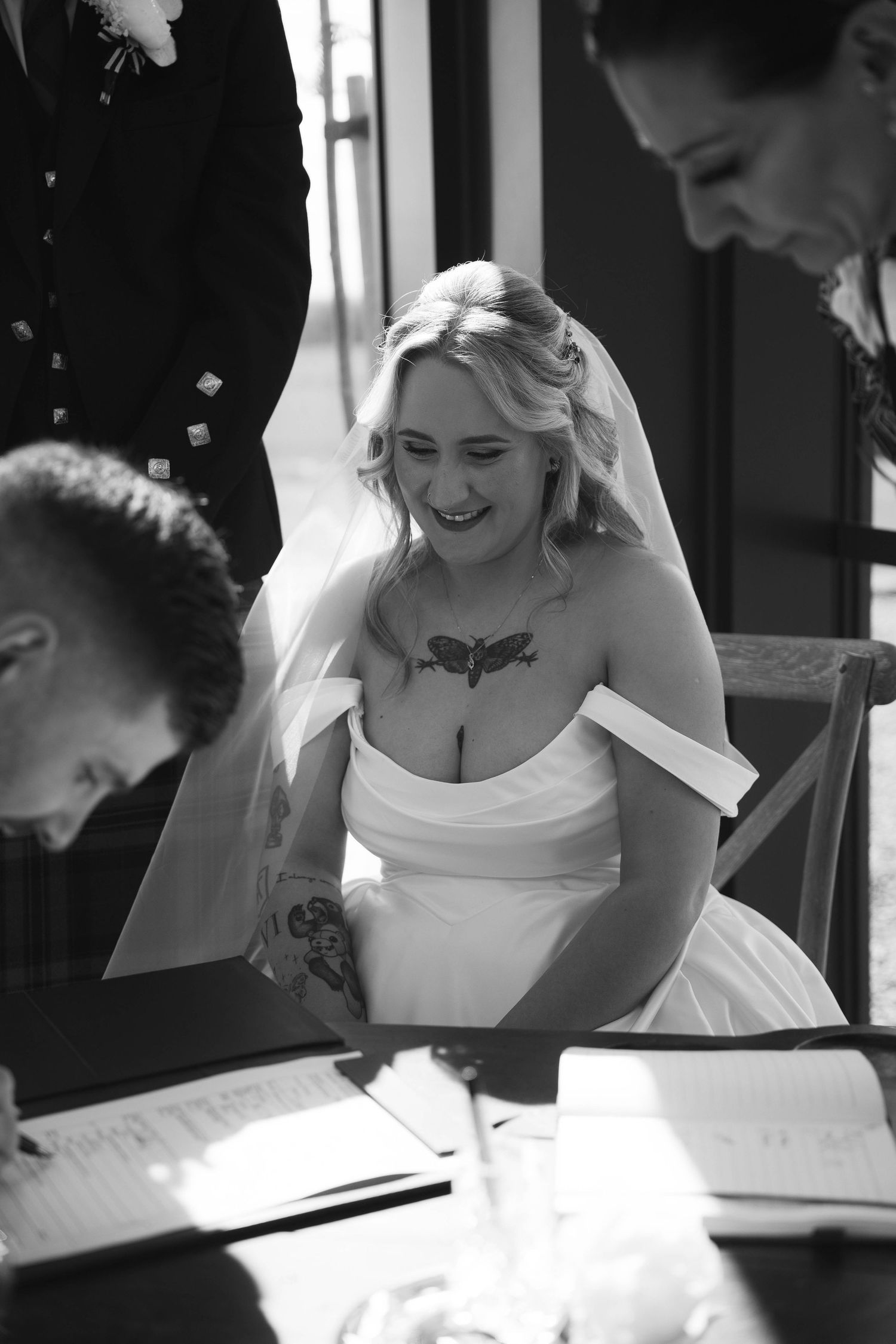 Tattooed bride in white gown and veil laughs joyfully while signing wedding register at ceremony.