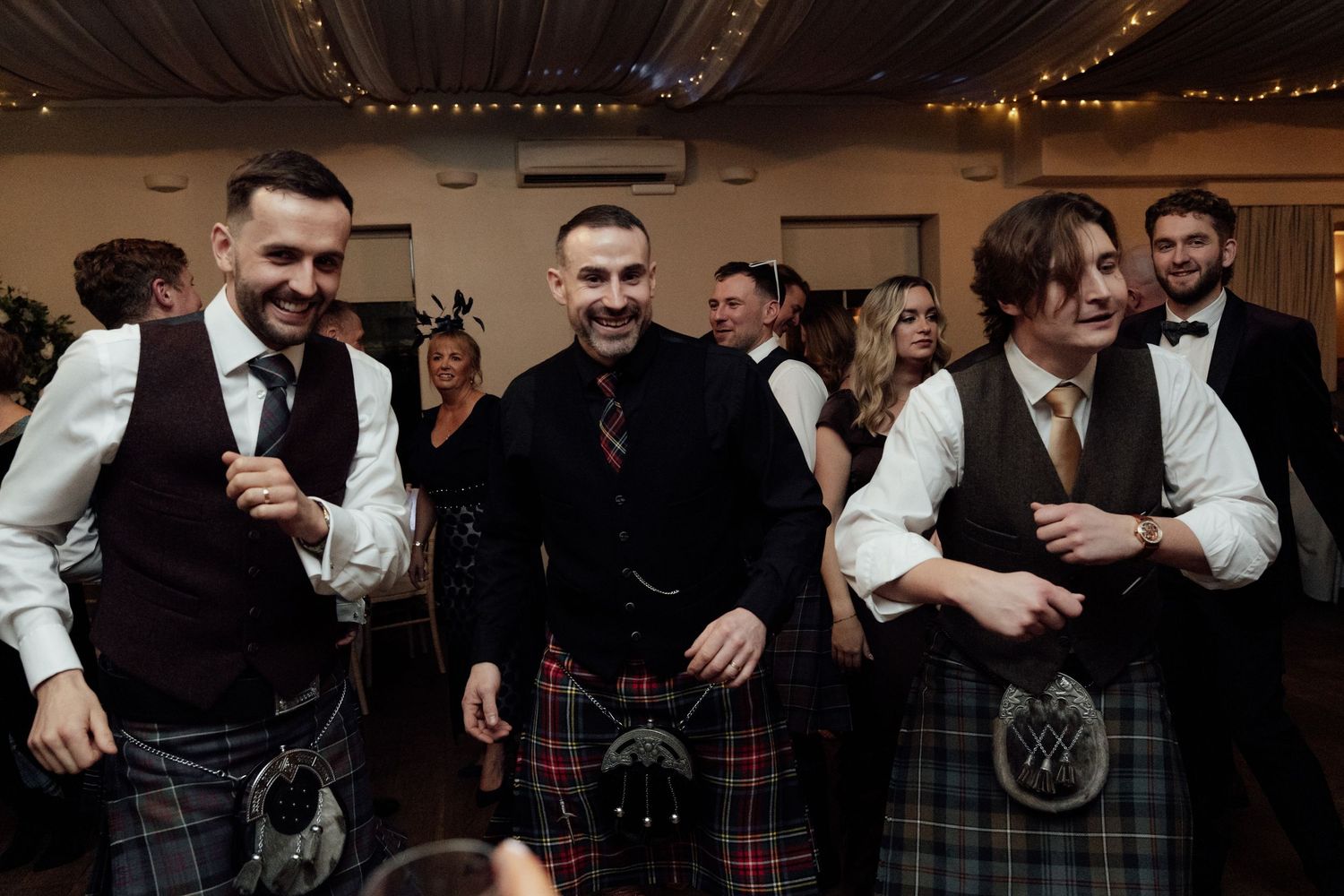 Wedding party members in kilts and formal attire celebrate during an energetic reception.