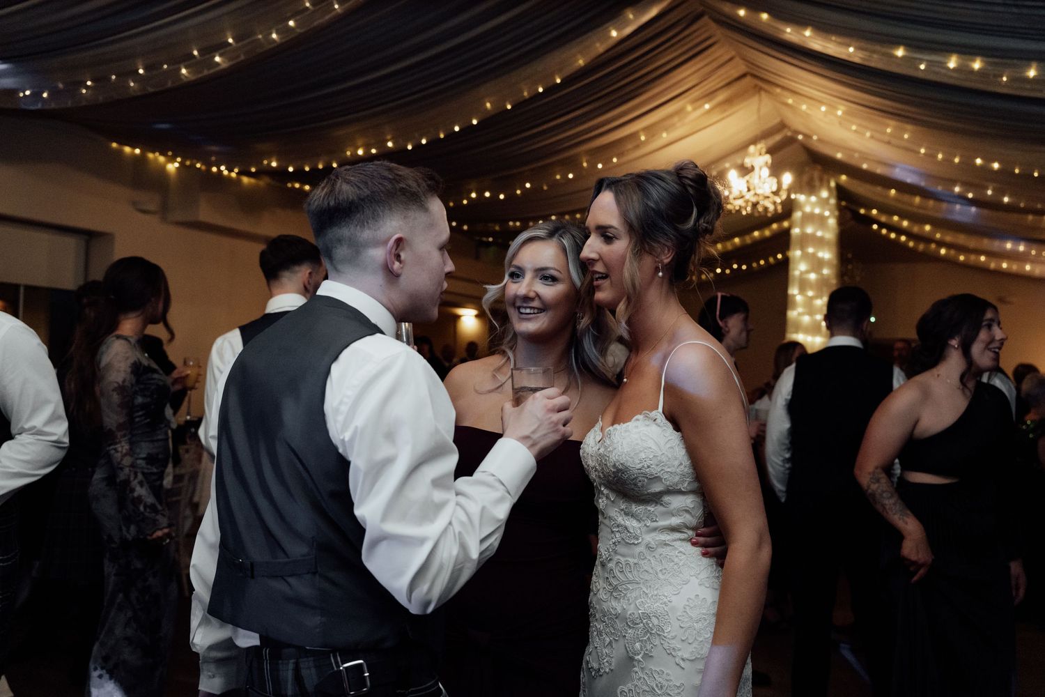 Wedding celebration with guests dancing and laughing under twinkling lights and draped fabric ceiling.
