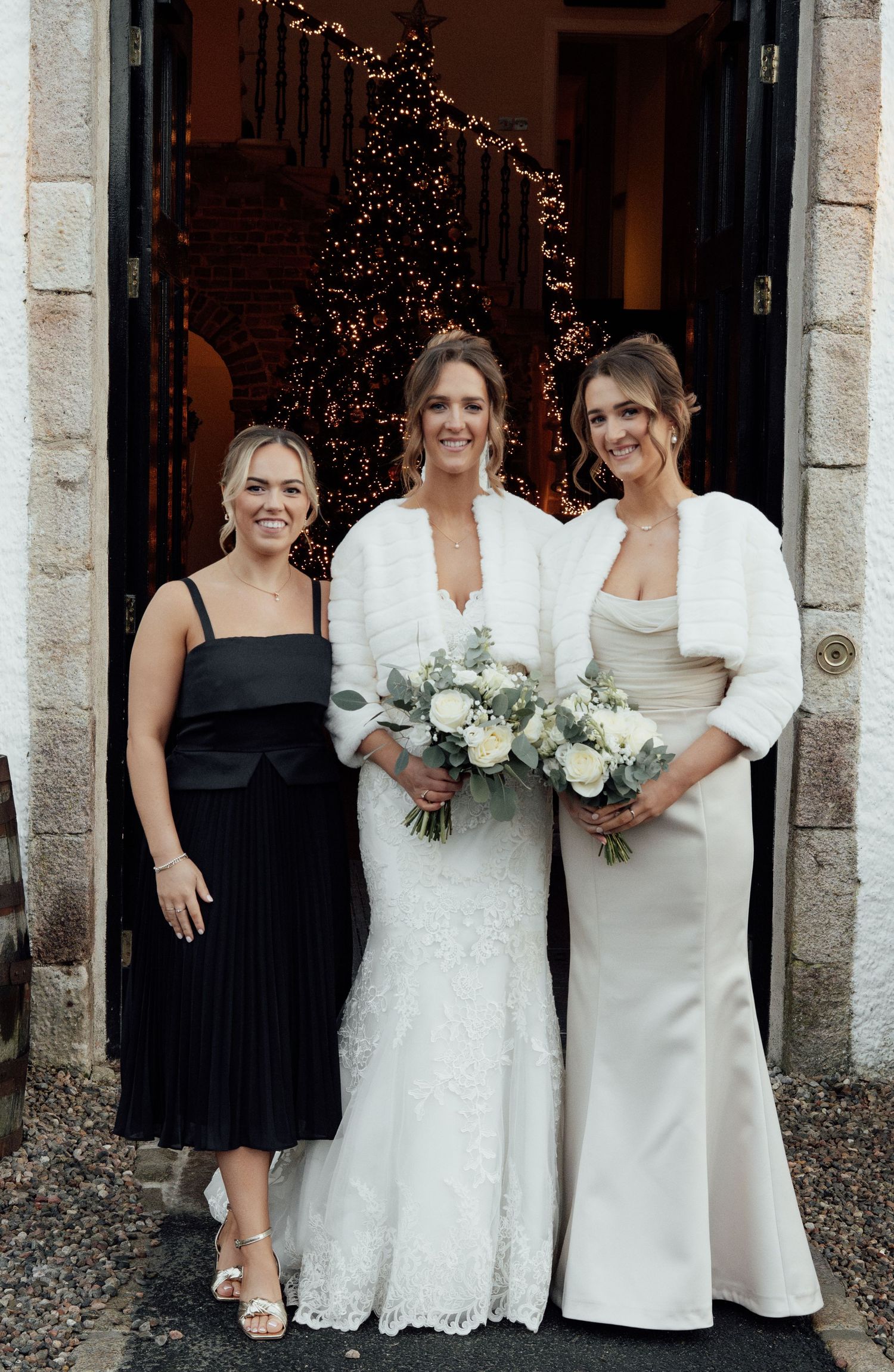 Wedding group portrait with bridesmaids in black dresses and white gowns standing by decorated doorway.
