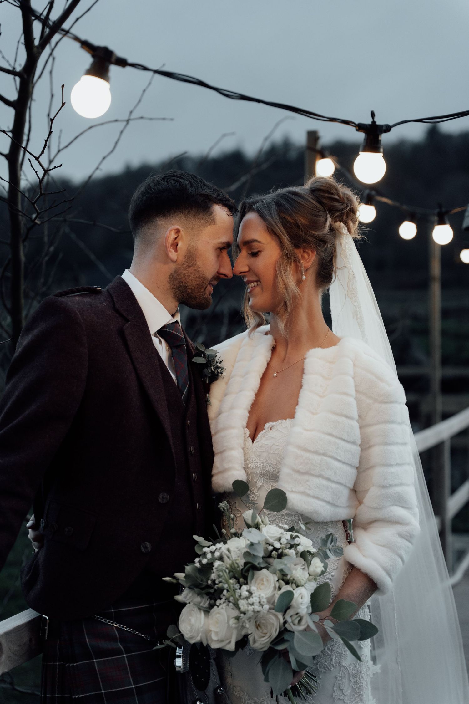 Intimate wedding portrait on bridge with string lights creating romantic atmosphere.