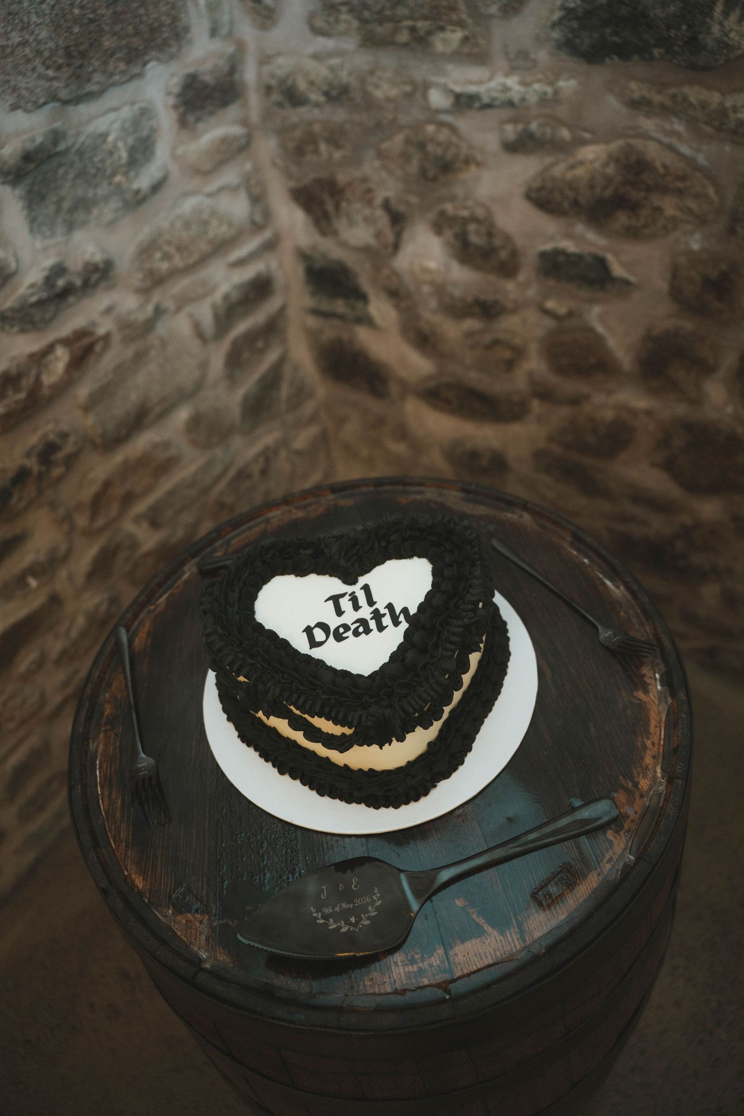 Heart-shaped chocolate wedding cake inscribed Til Death displayed on rustic wooden barrel against stone wall.