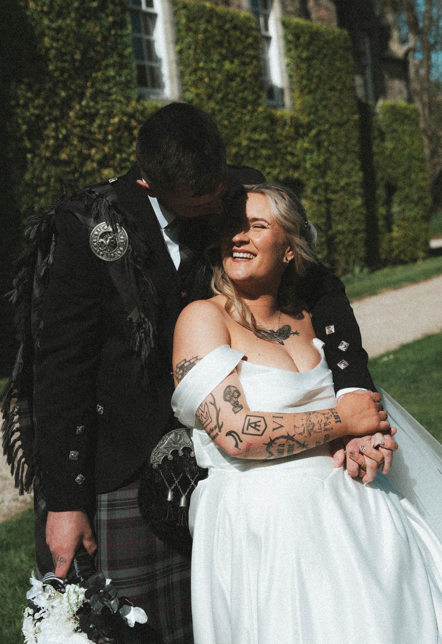 Laughing tattooed bride in white gown embraced by groom in Scottish kilt in sunny garden setting.