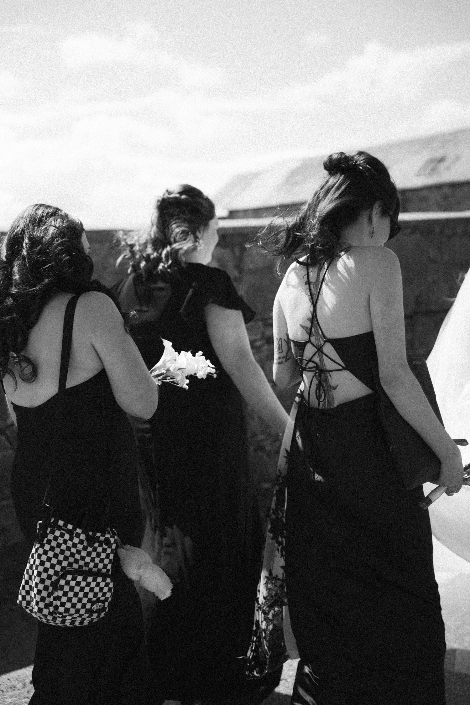 Three bridesmaids in black dresses walking outdoors, one carrying white flowers, captured in black and white.