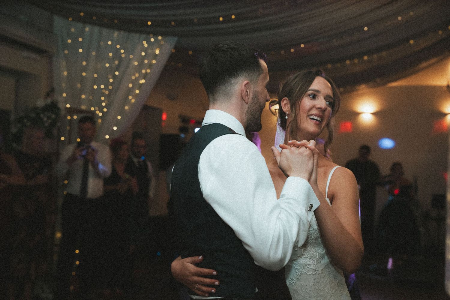 Couples dancing at a wedding reception under fairy lights and draped ceiling decor.