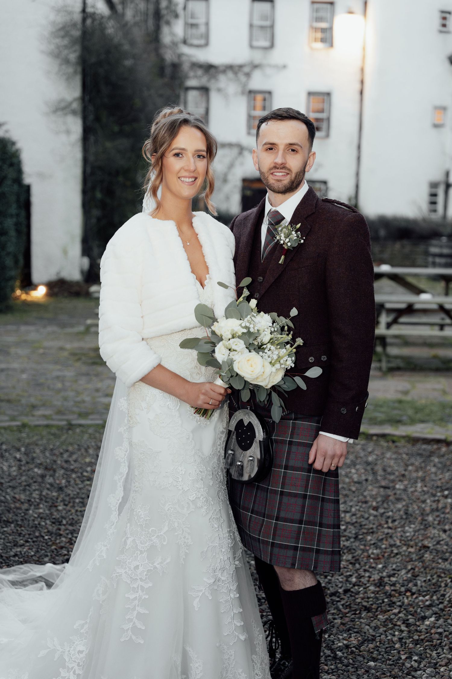 Wedding couple poses in front of white building, bride in lace gown and groom wearing traditional Scottish kilt.
