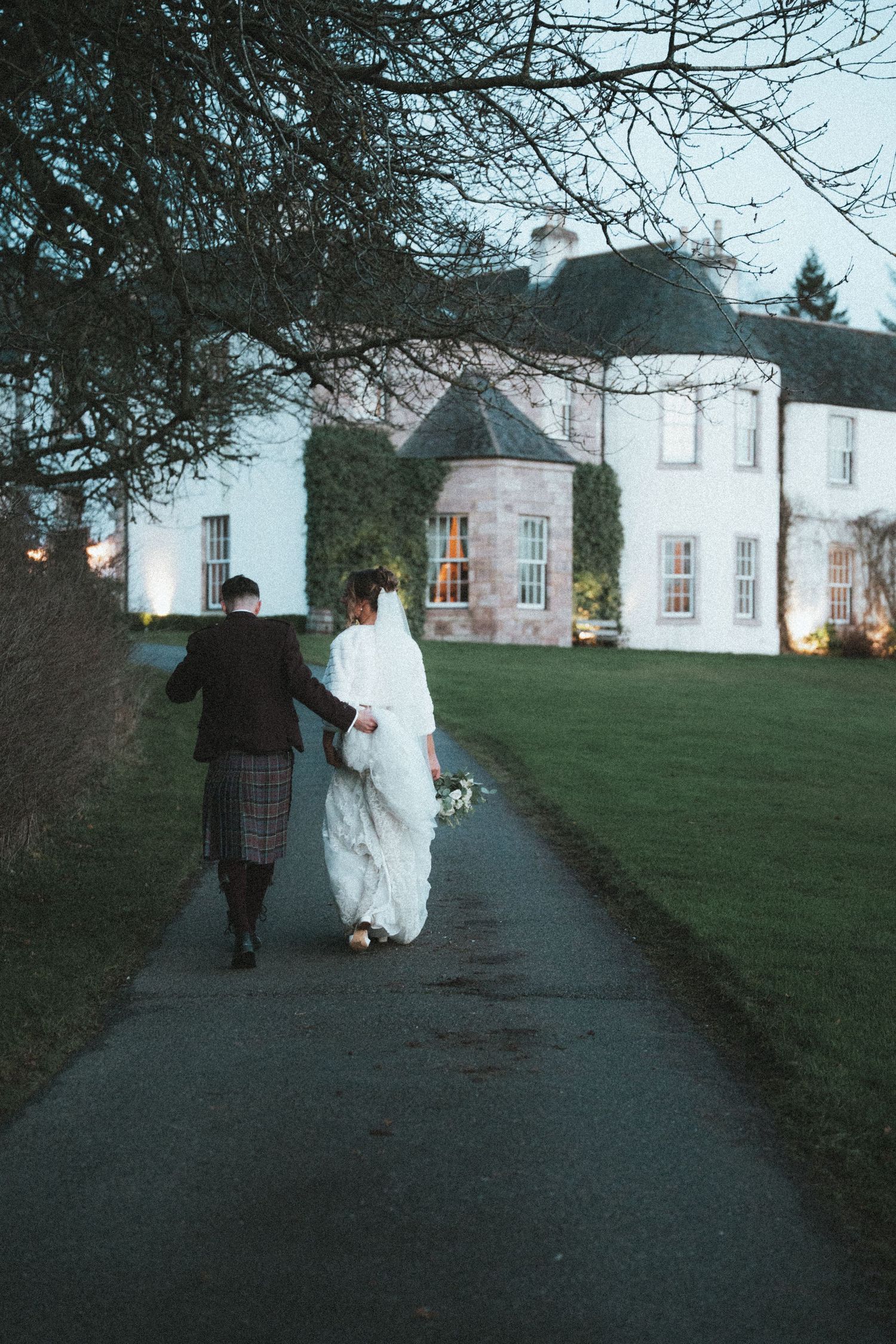 Wedding couple playfully runs along a path towards a grand white building at dusk.