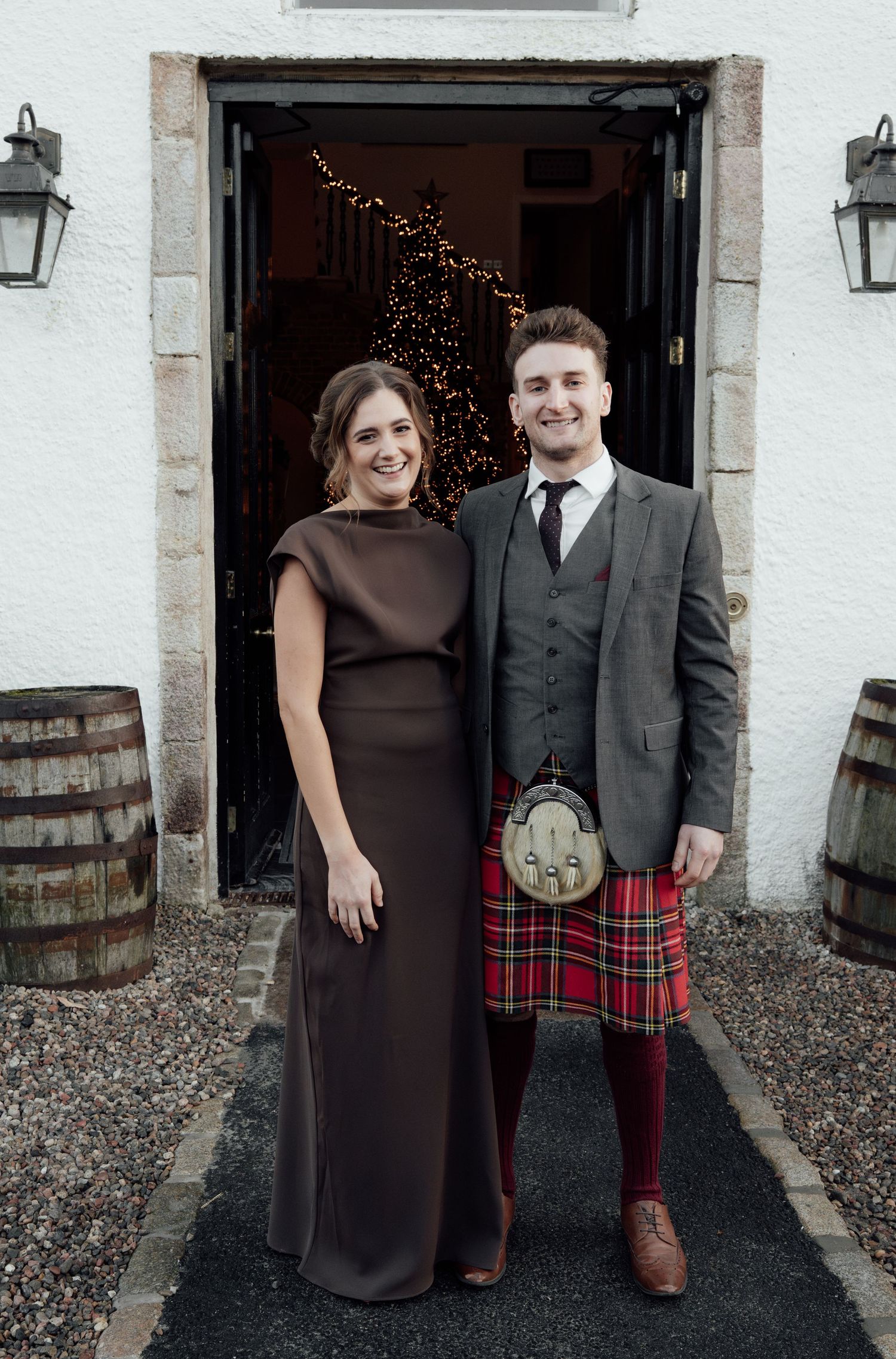 A formal portrait of a couple standing in a doorway with fairy lights, featuring Scottish formal wear.