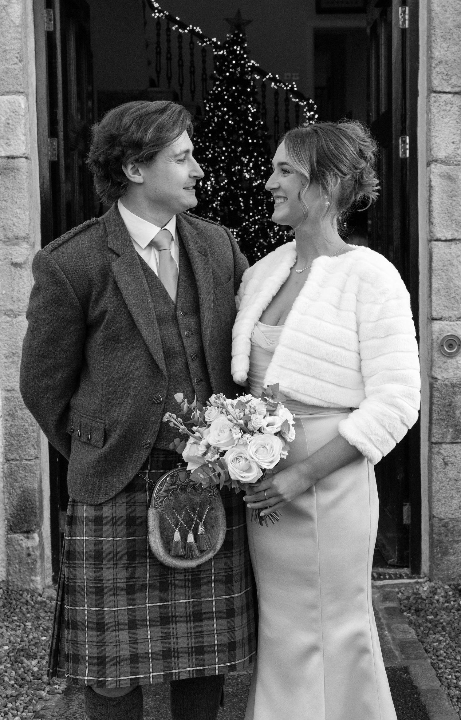 A couple in wedding attire share a tender moment outside a stone building with the groom wearing a traditional kilt.