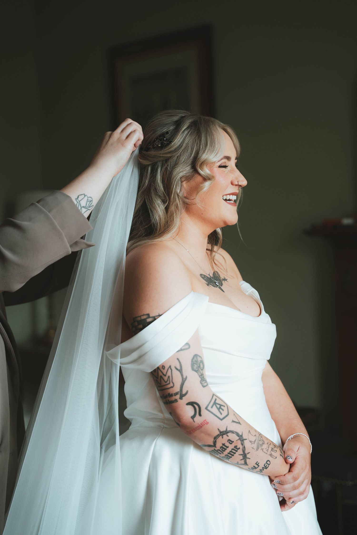 Smiling bride with tattoos in off-shoulder white gown having veil placed during wedding preparations.