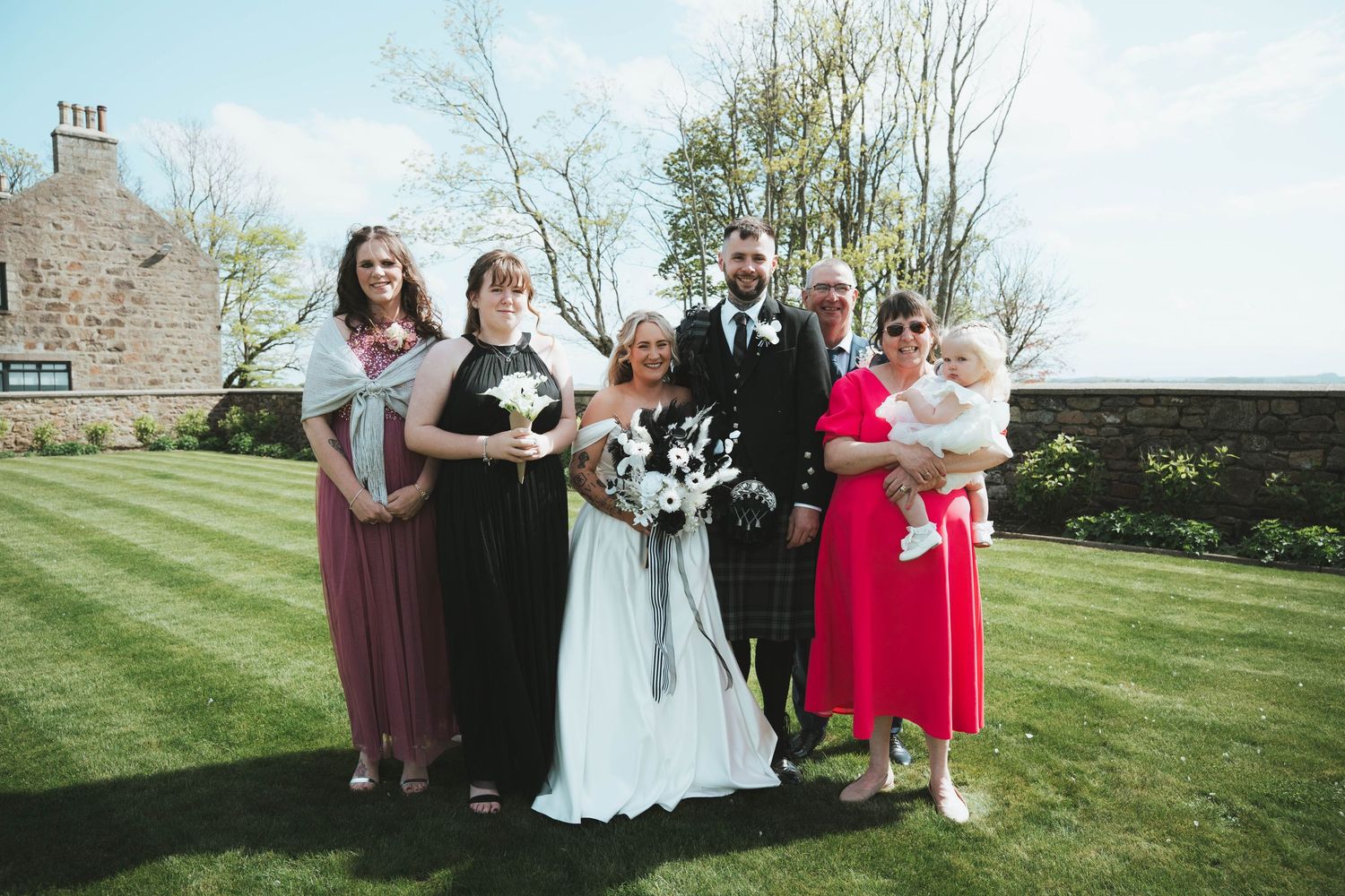 Wedding party group portrait on sunny lawn with bride in white gown holding black and white bouquet.
