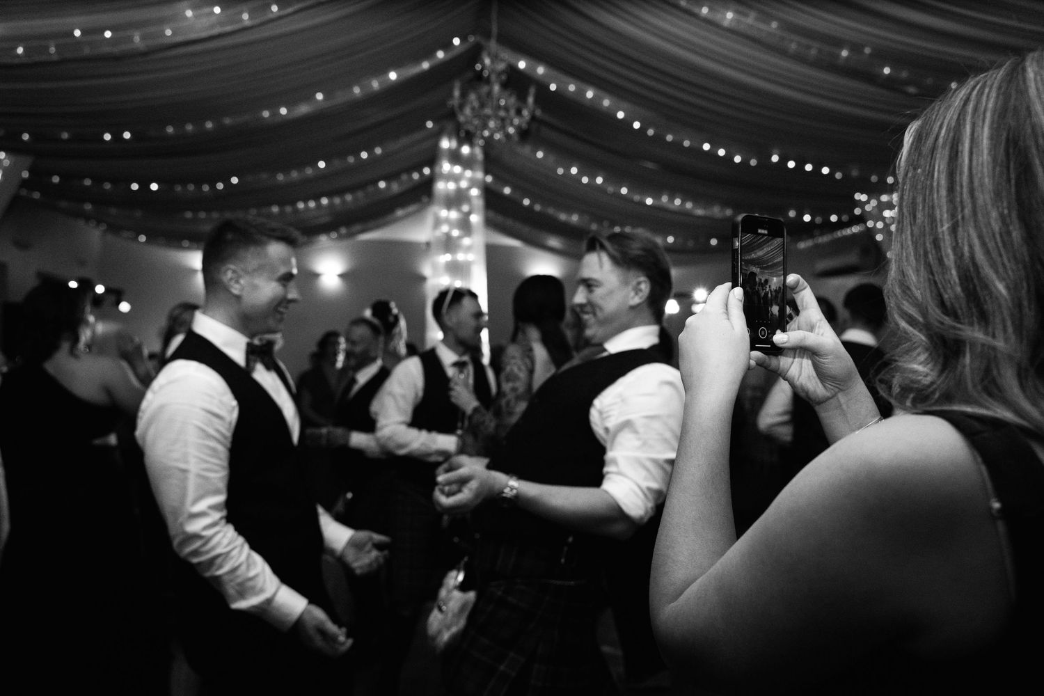 Black and white photo of guests dancing at a wedding reception with string lights overhead.