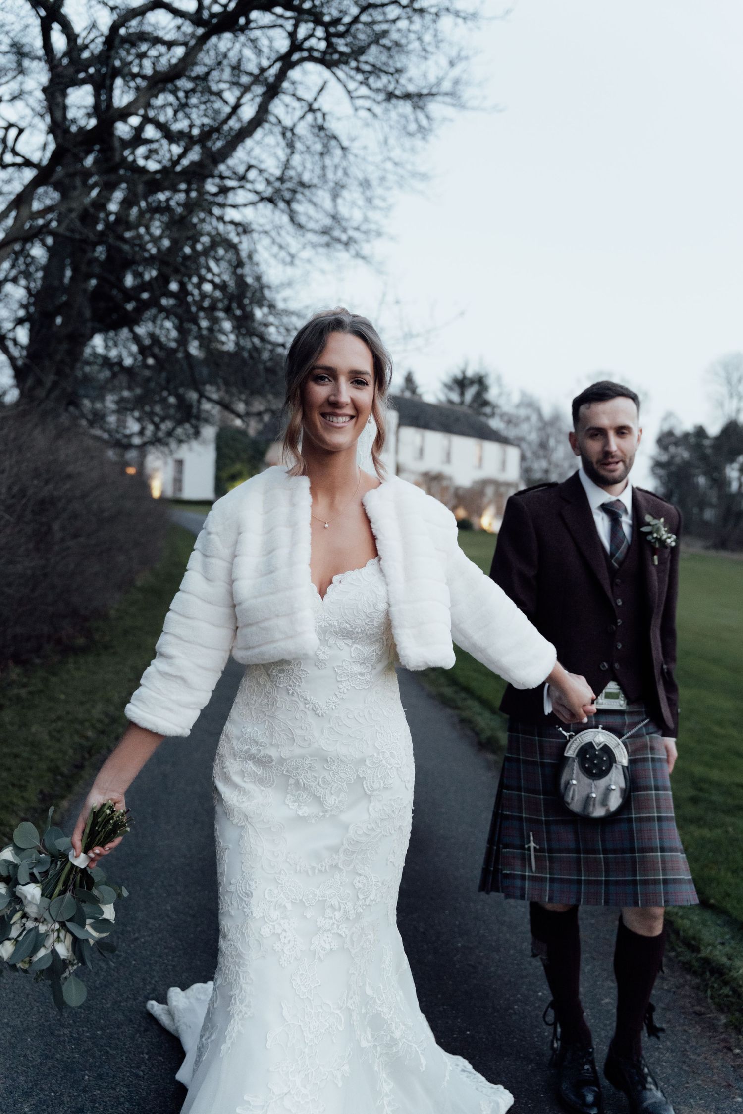 Wedding couple walks along a winter path, bride in white dress and groom wearing traditional Scottish attire.