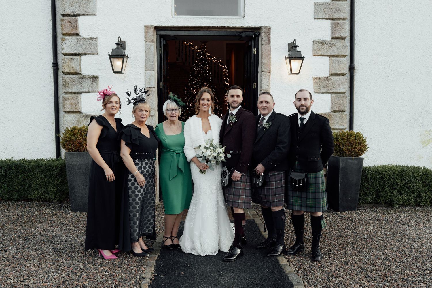 Wedding party members in kilts and formal attire gather for photos at venue entrance.