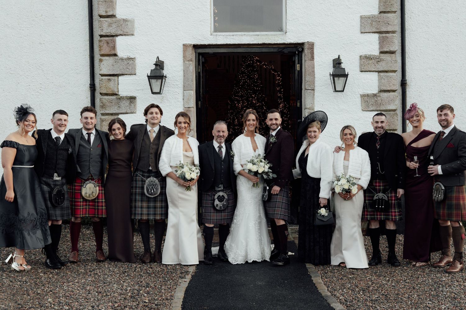 Wedding celebration outside historic stone building with Scottish kilts and formal wear visible in group photo.
