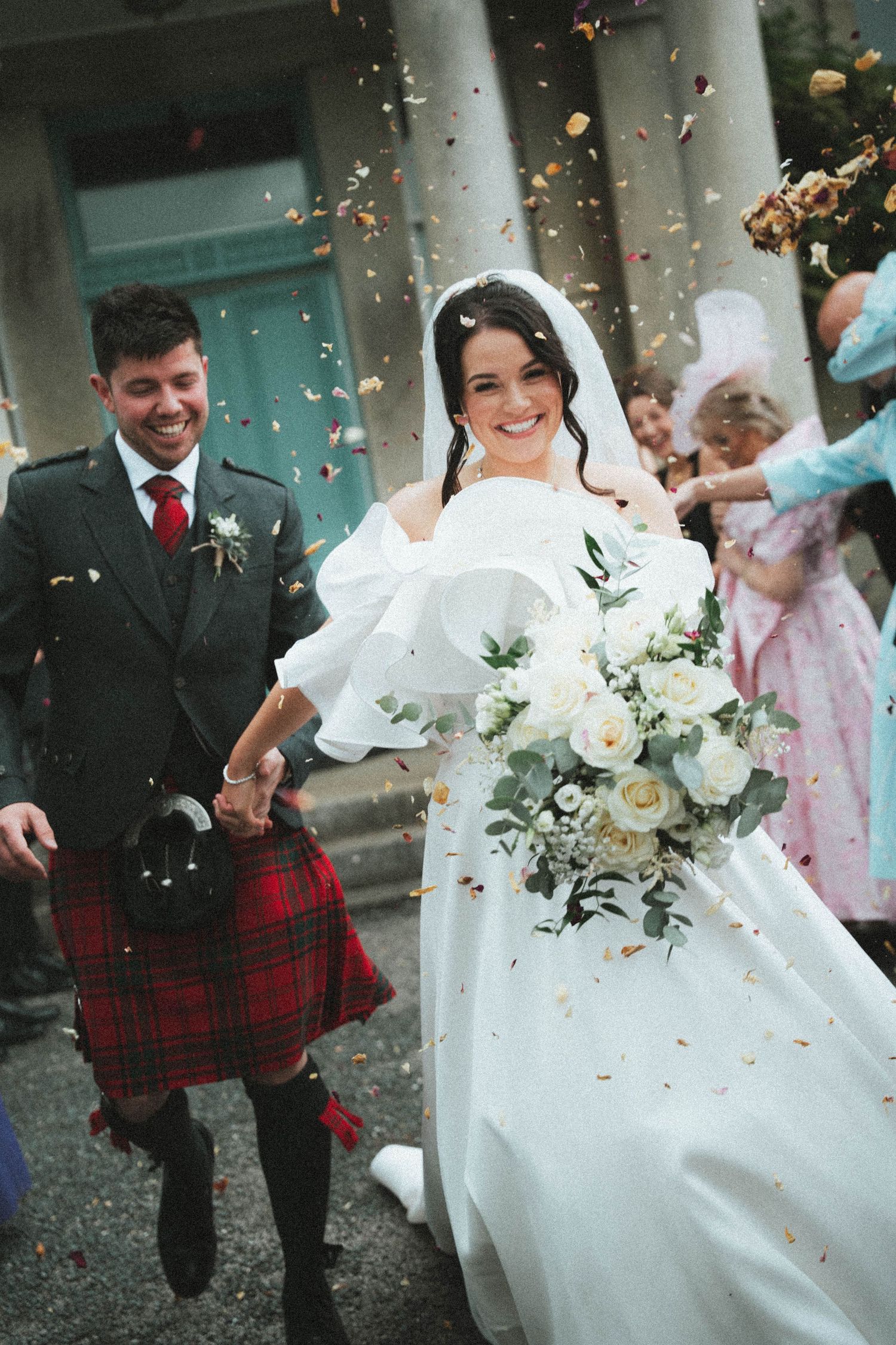 Bride and groom walk through confetti after Scottish wedding ceremony, groom wearing red tartan kilt, bride holding white floral bouquet.