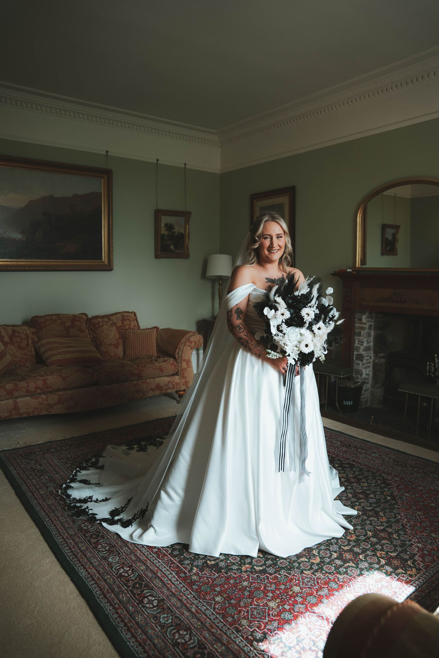 Elegant bride in off-shoulder satin wedding gown holding white floral bouquet standing in a classic decorated room.