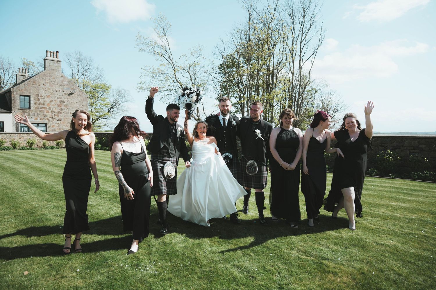 Wedding party celebrating outdoors on green lawn, bride in white gown with groomsmen in kilts and bridesmaids in black dresses.