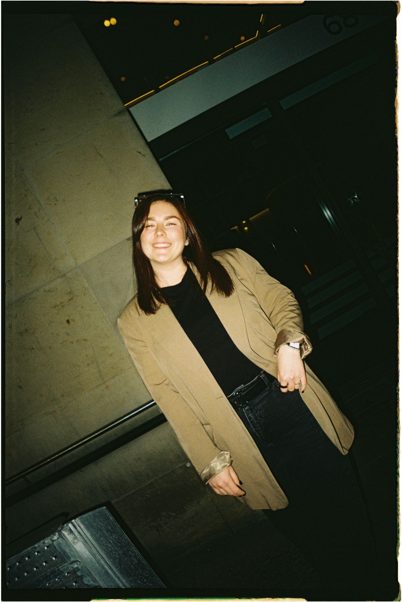 A person in a tan coat and black outfit stands in a dark hallway.