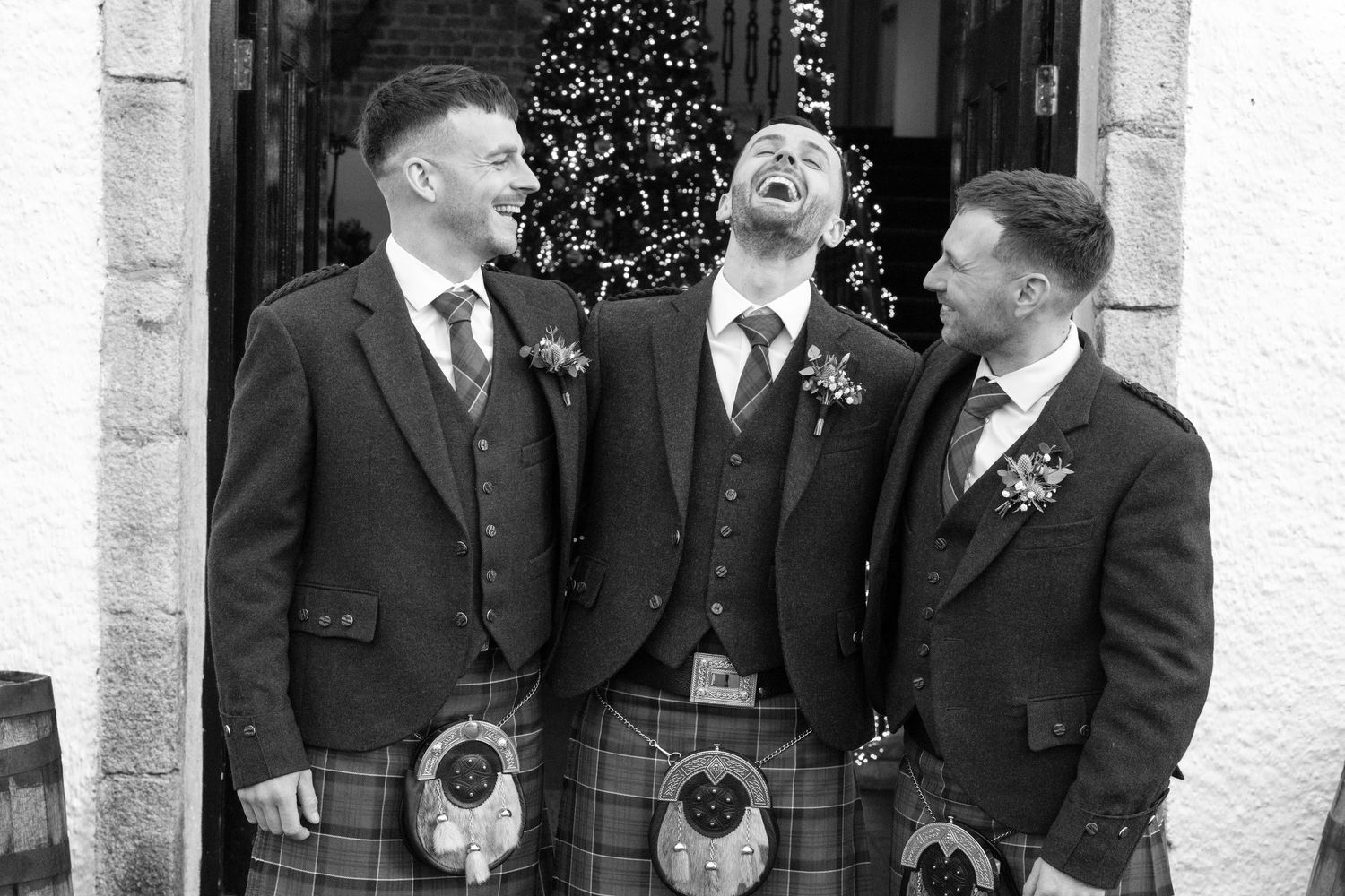 Black and white photo of three groomsmen in kilts sharing a laugh at church entrance.