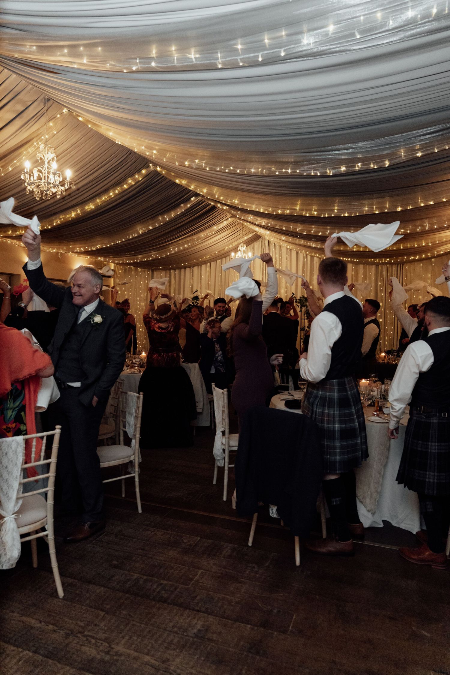 Wedding reception venue decorated with white draping and string lights as guests mingle on dance floor.