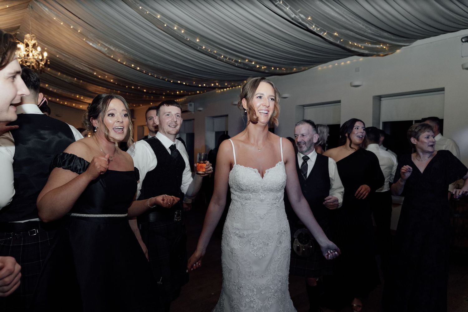 Wedding reception guests dancing and celebrating on a dimly lit dance floor under draped fabric ceiling.