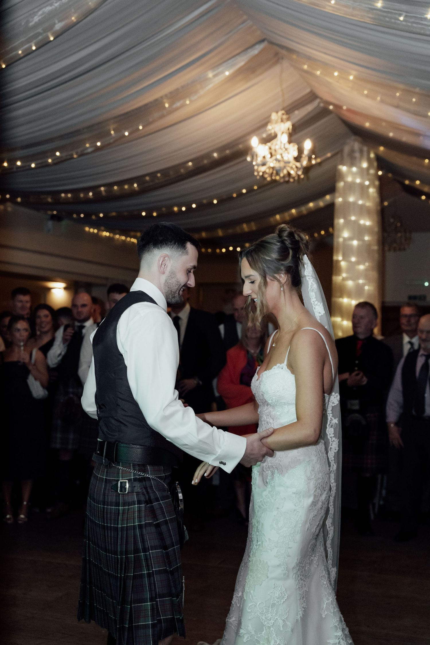Bride in white dress dances with partner wearing traditional Scottish kilt at reception.