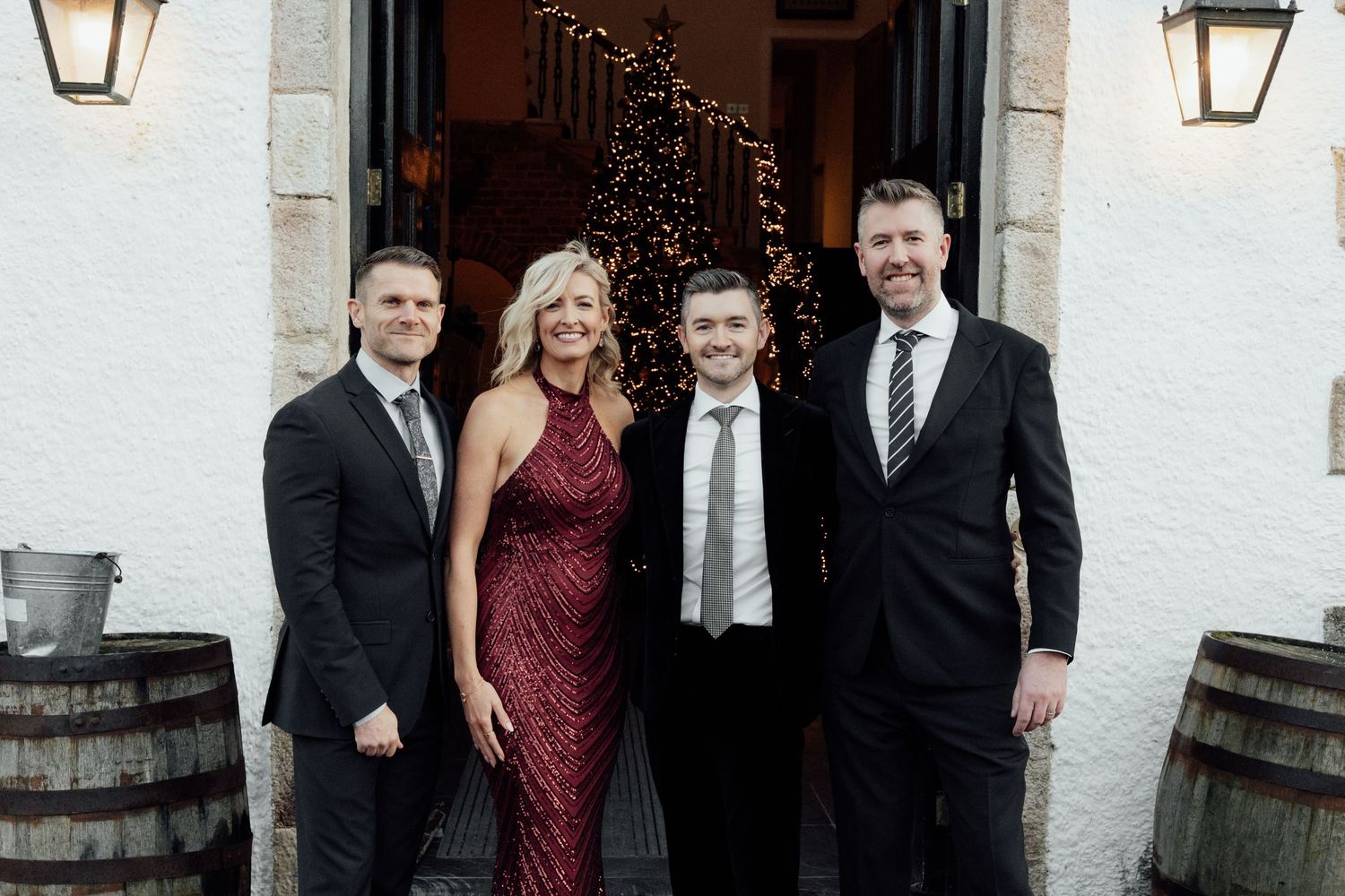 Formal group photo of wedding attendees outside white stone building with decorative lighting.