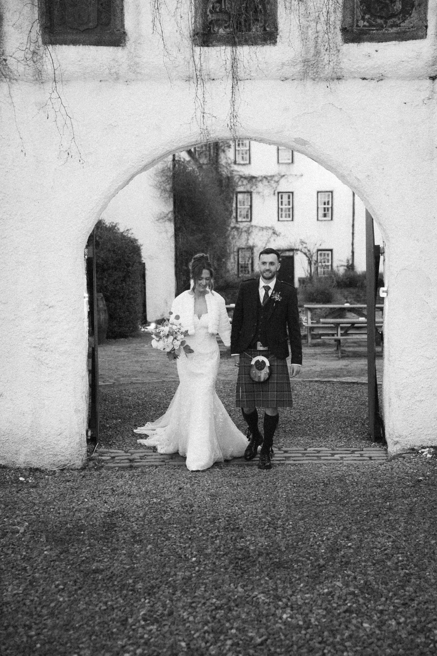 Black and white wedding photo under stone archway with couple in traditional Scottish wedding attire.