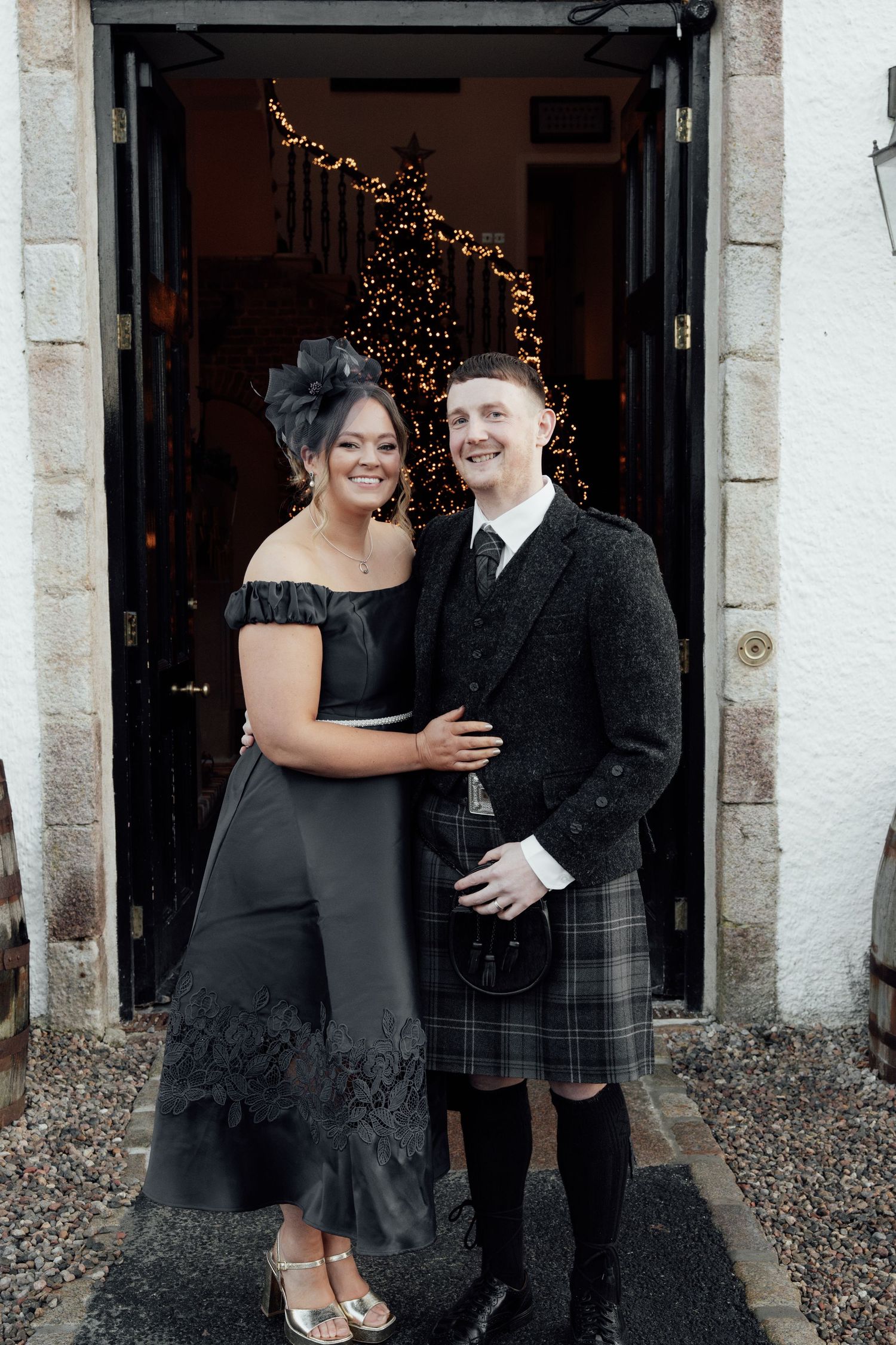 Wedding party members in black dresses and kilts share laughs outside a venue with fairy lights.