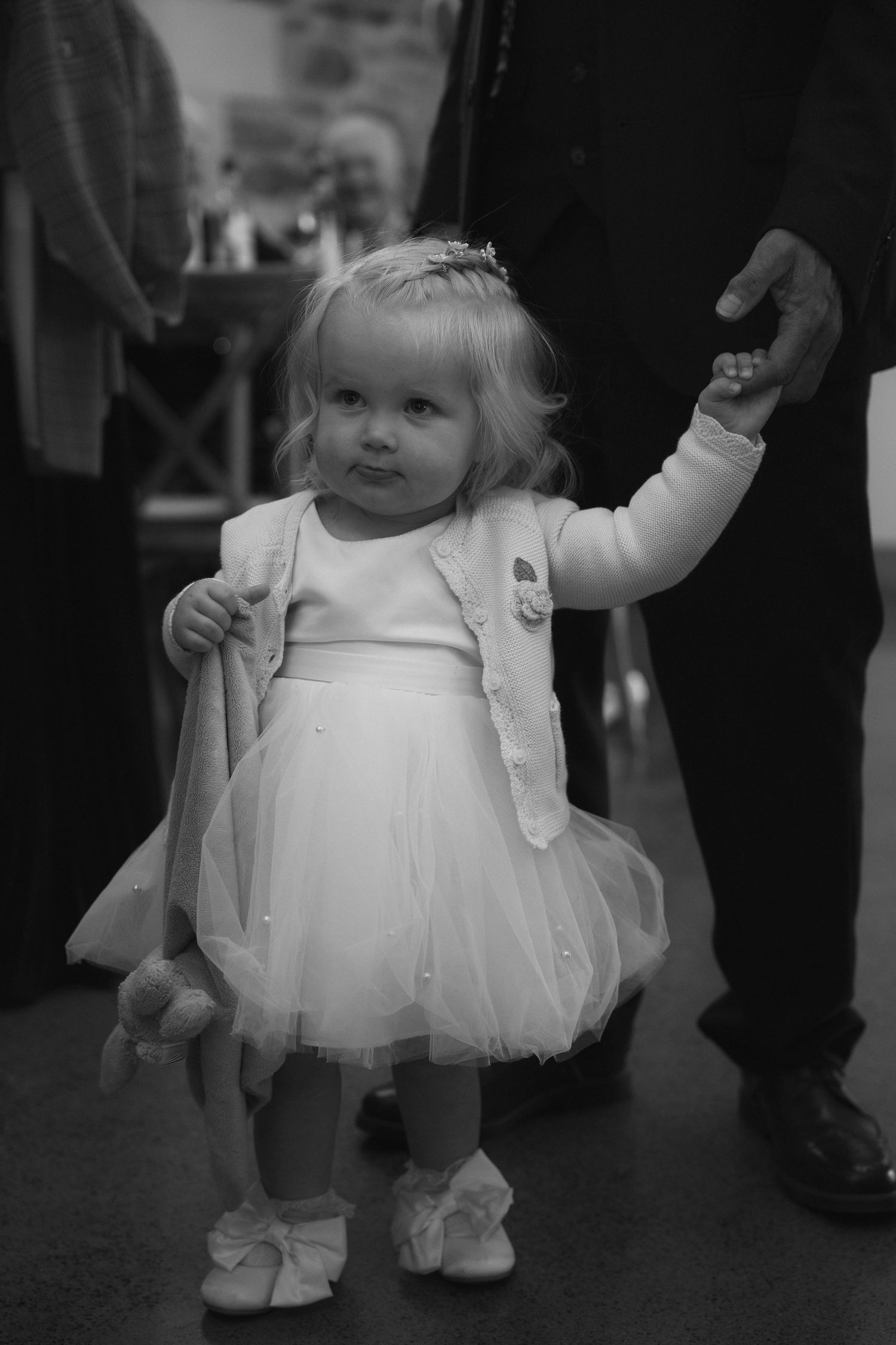 Sweet toddler flower girl in white tutu dress holding an adults hand at a wedding reception.