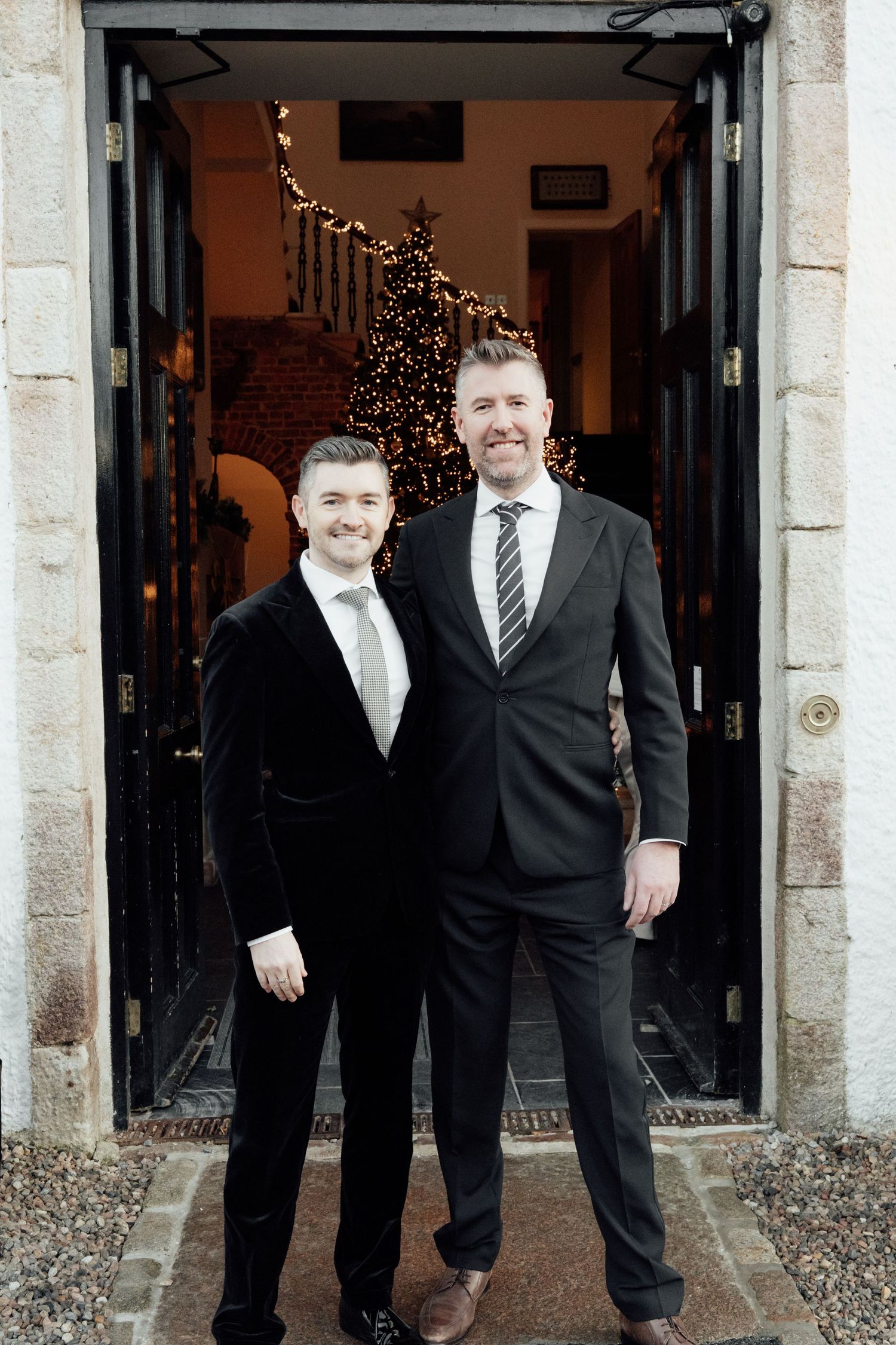 Two wedding guests pose in dark suits at stone building entrance with holiday lights visible.