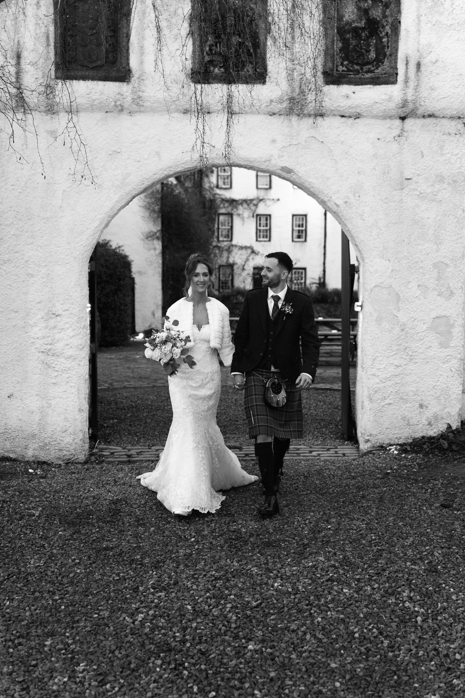 Black and white portrait of newlyweds walking through stone archway in Scottish wedding attire.