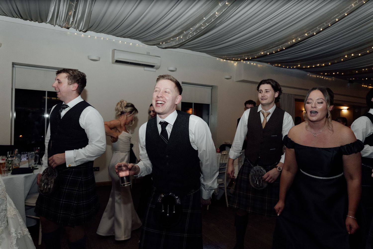Wedding celebration showing guests dancing in a decorated venue with fabric-draped ceiling.