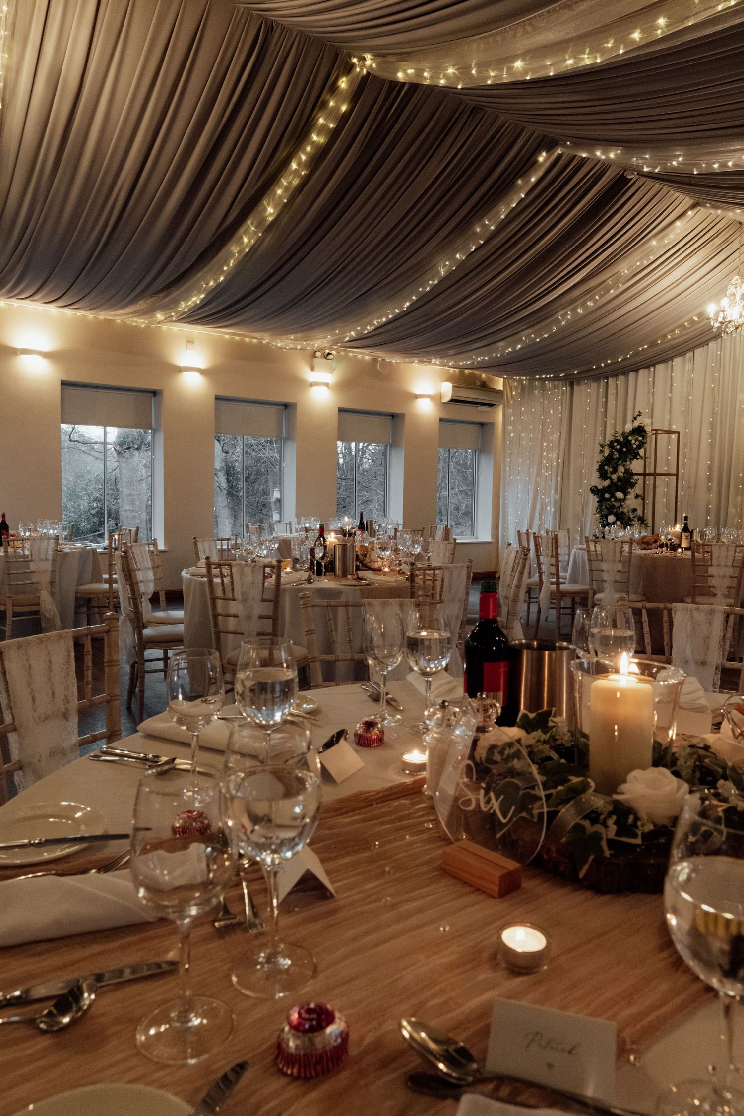 Wedding reception room with fabric-draped ceiling, fairy lights and decorated round tables.