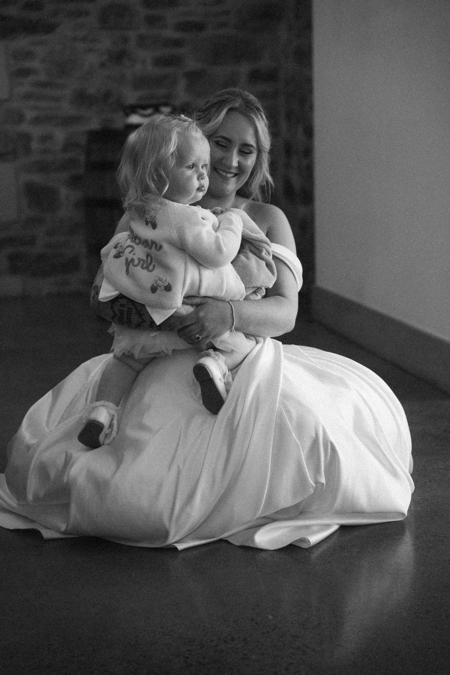 Bride in white gown sits on floor smiling while holding a toddler in black and white photo.