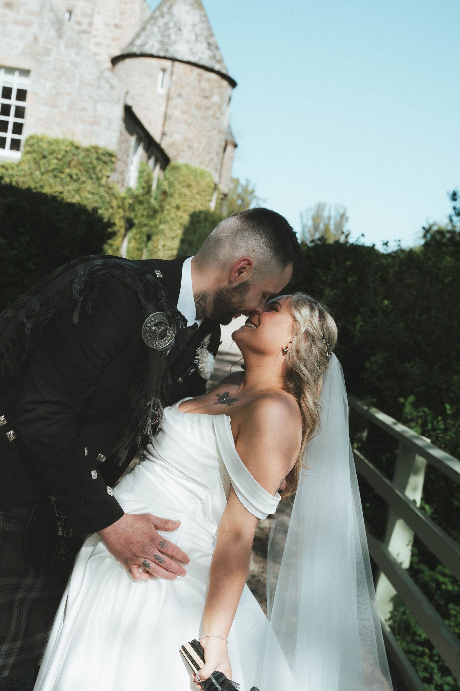 A bride and groom share a romantic kiss outside a historic stone castle, with the bride wearing a white gown and veil.