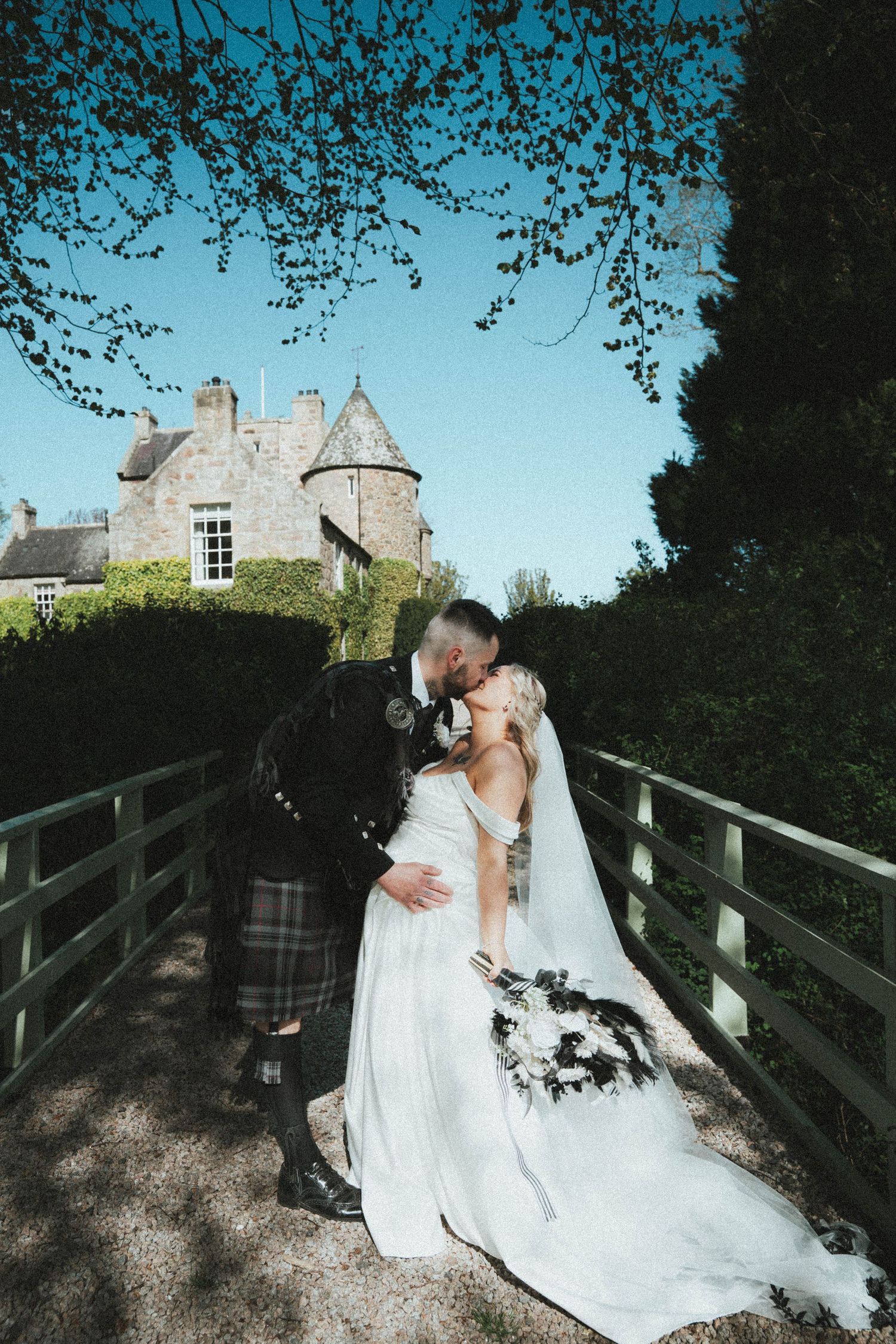 Bride and groom kissing on bridge before Scottish castle surrounded by lush greenery on sunny day.