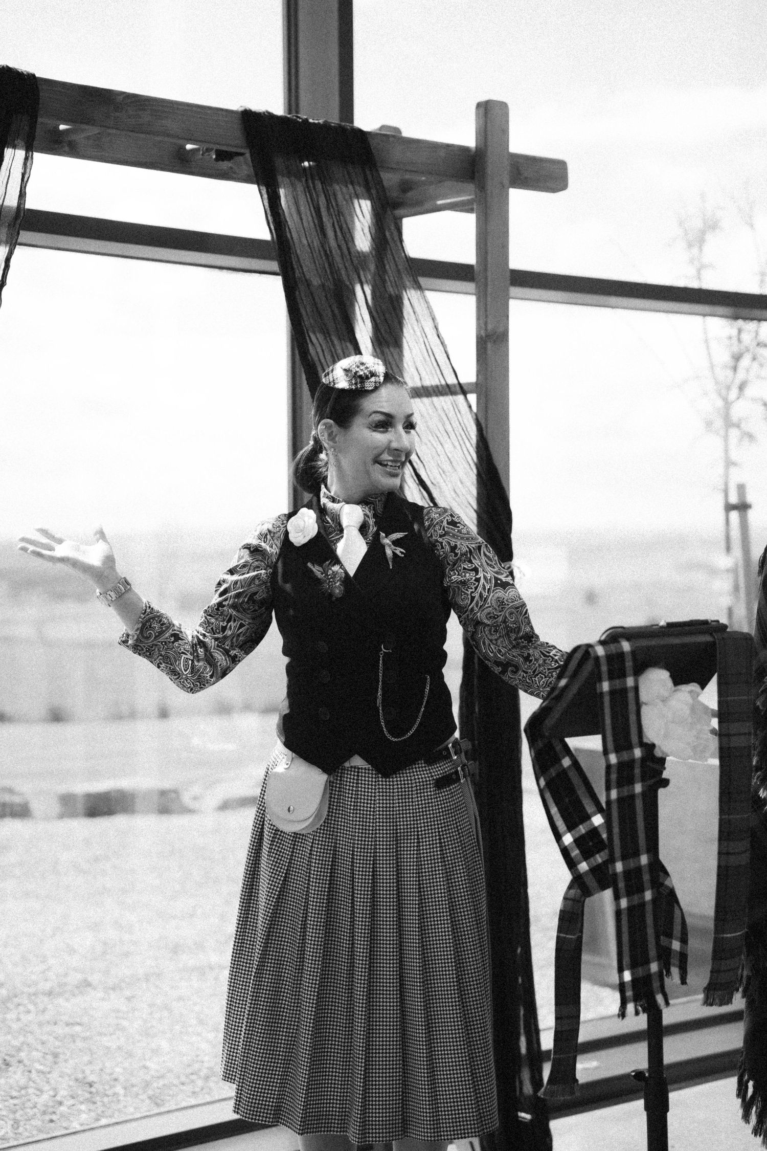 Woman in vintage Victorian-style outfit with vest and skirt gesturing welcomingly indoors in black and white.