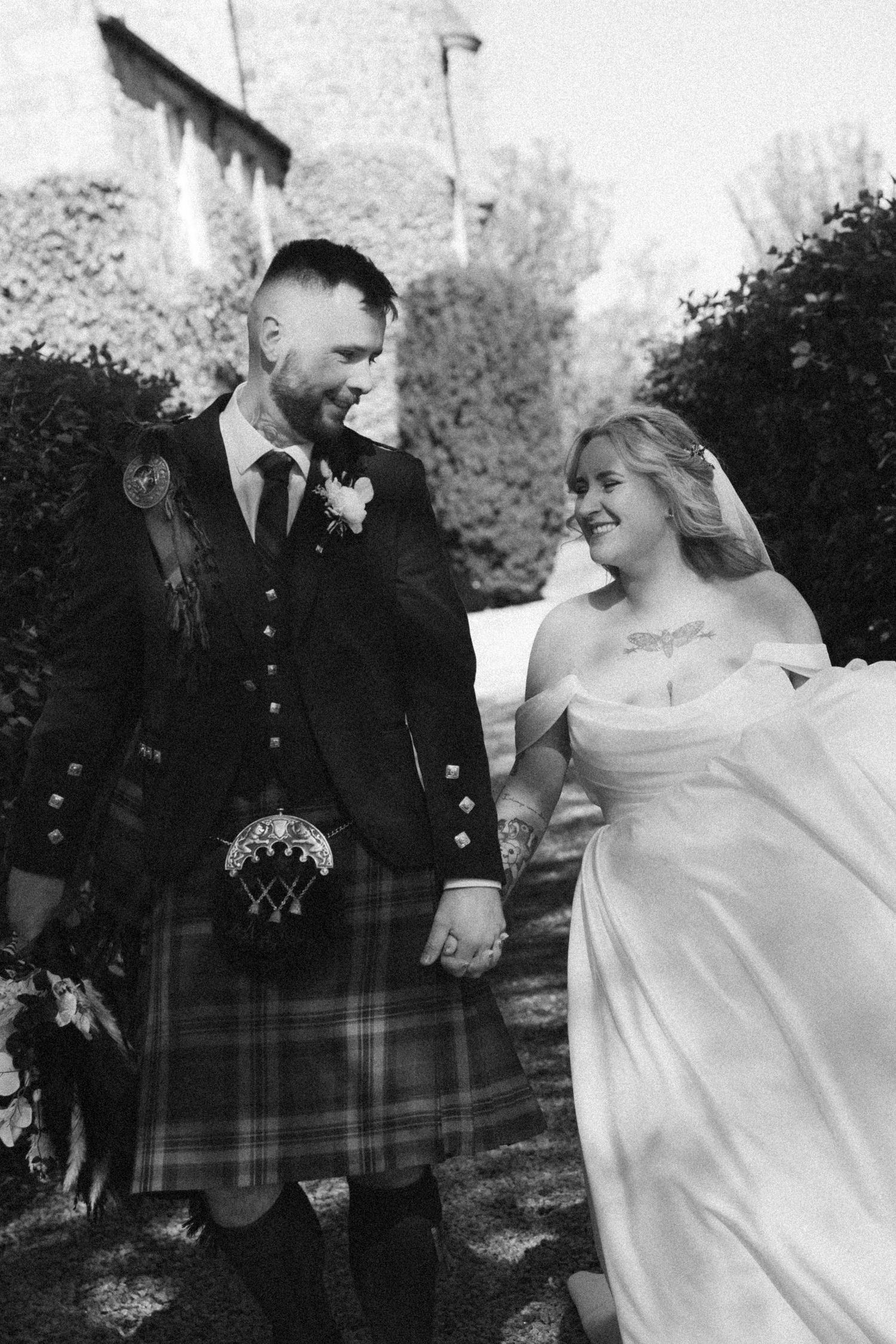 Black and white photo of smiling bride and groom in kilt walking hand in hand past a stone castle.