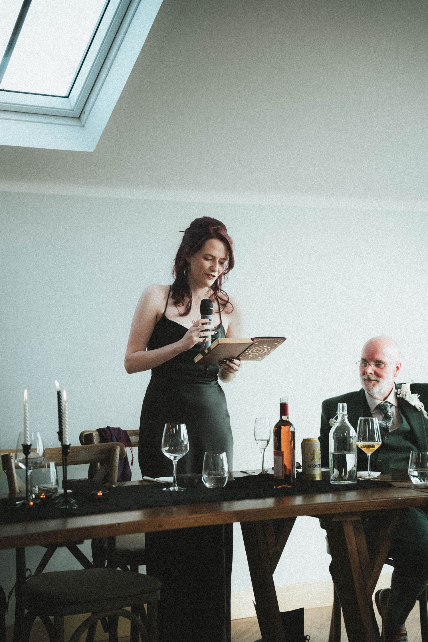 Woman in black dress giving speech at wedding reception holding microphone and notes at elegantly decorated dinner table.