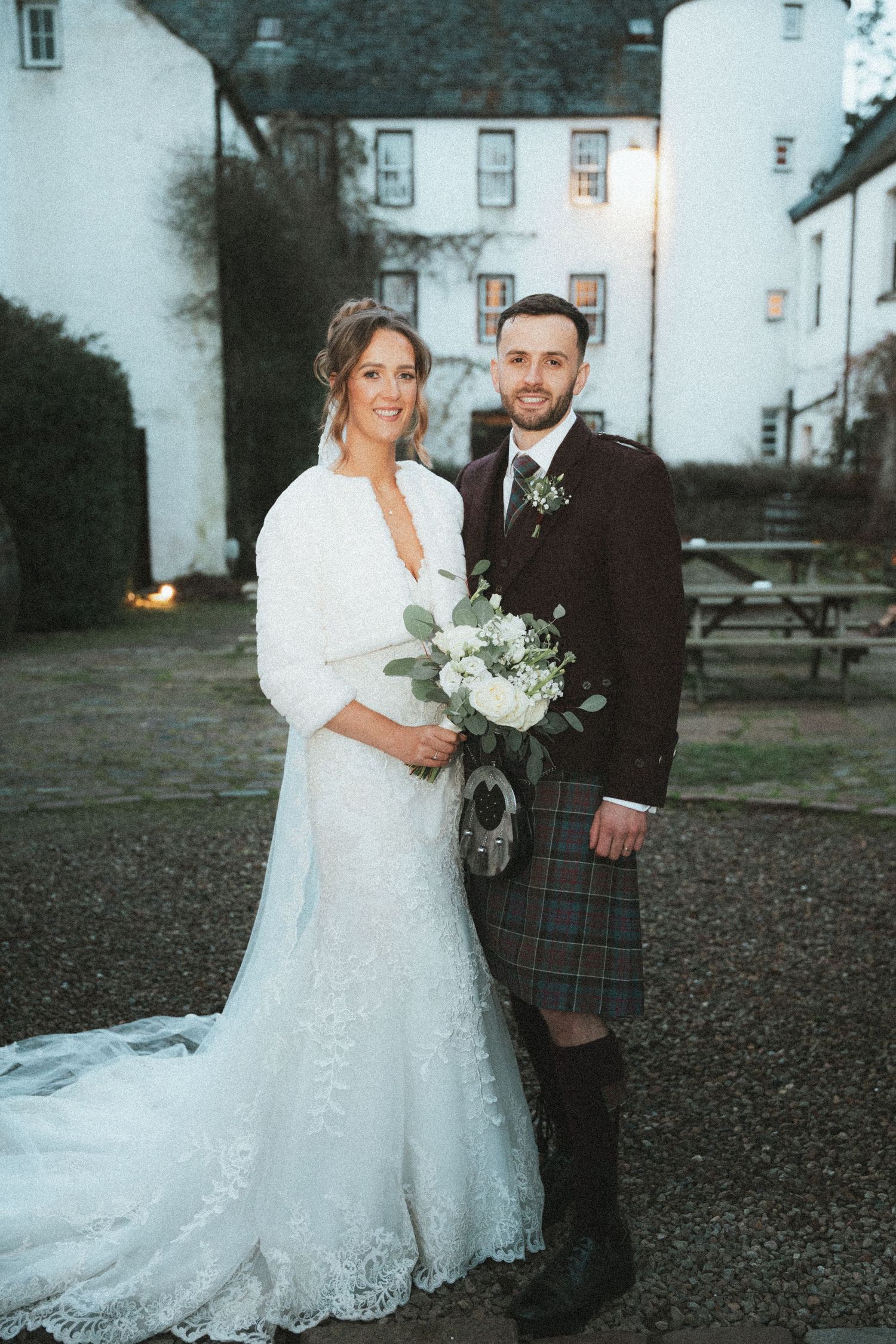 Elegant wedding portrait featuring couple in white dress and tartan kilt against historic Scottish architecture.