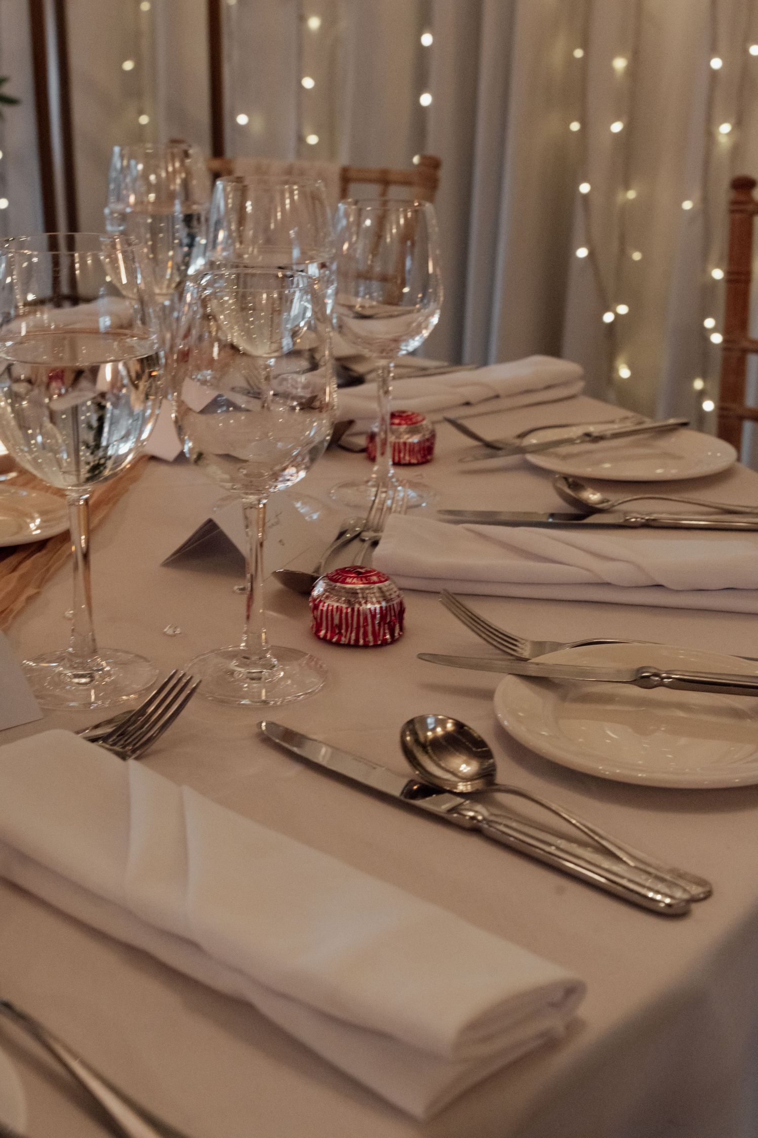 Elegant table setting with wine glasses, silverware and white linens illuminated by twinkle lights.