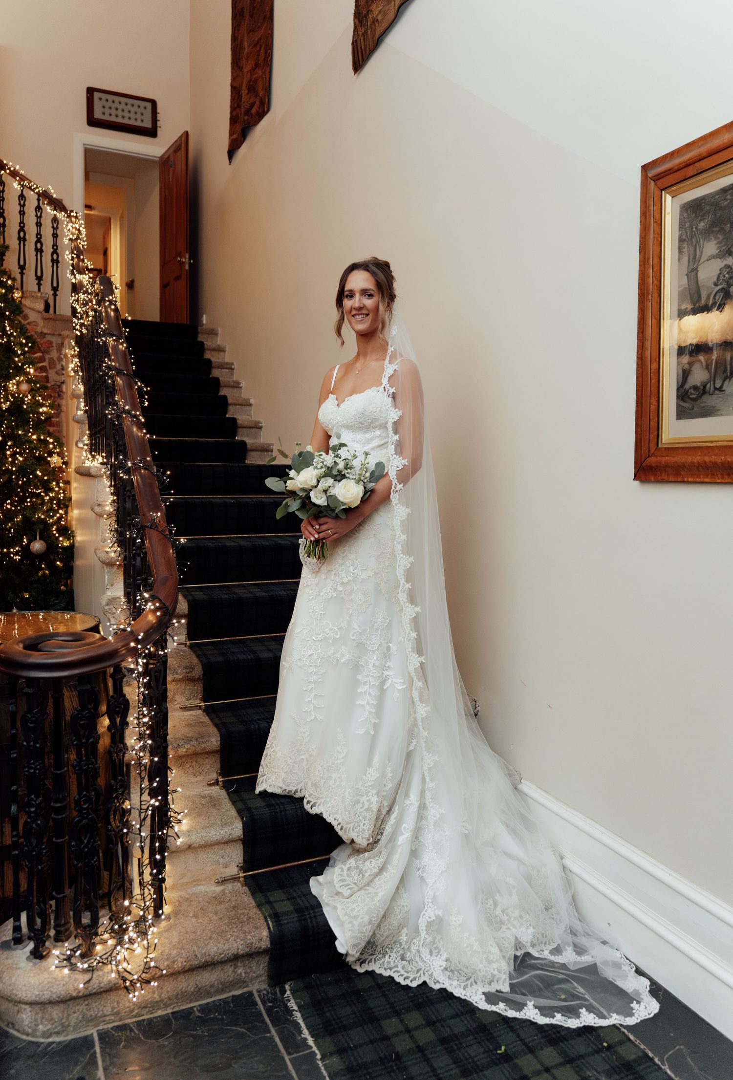 Elegant bride in lace wedding gown poses on staircase decorated with twinkling fairy lights.