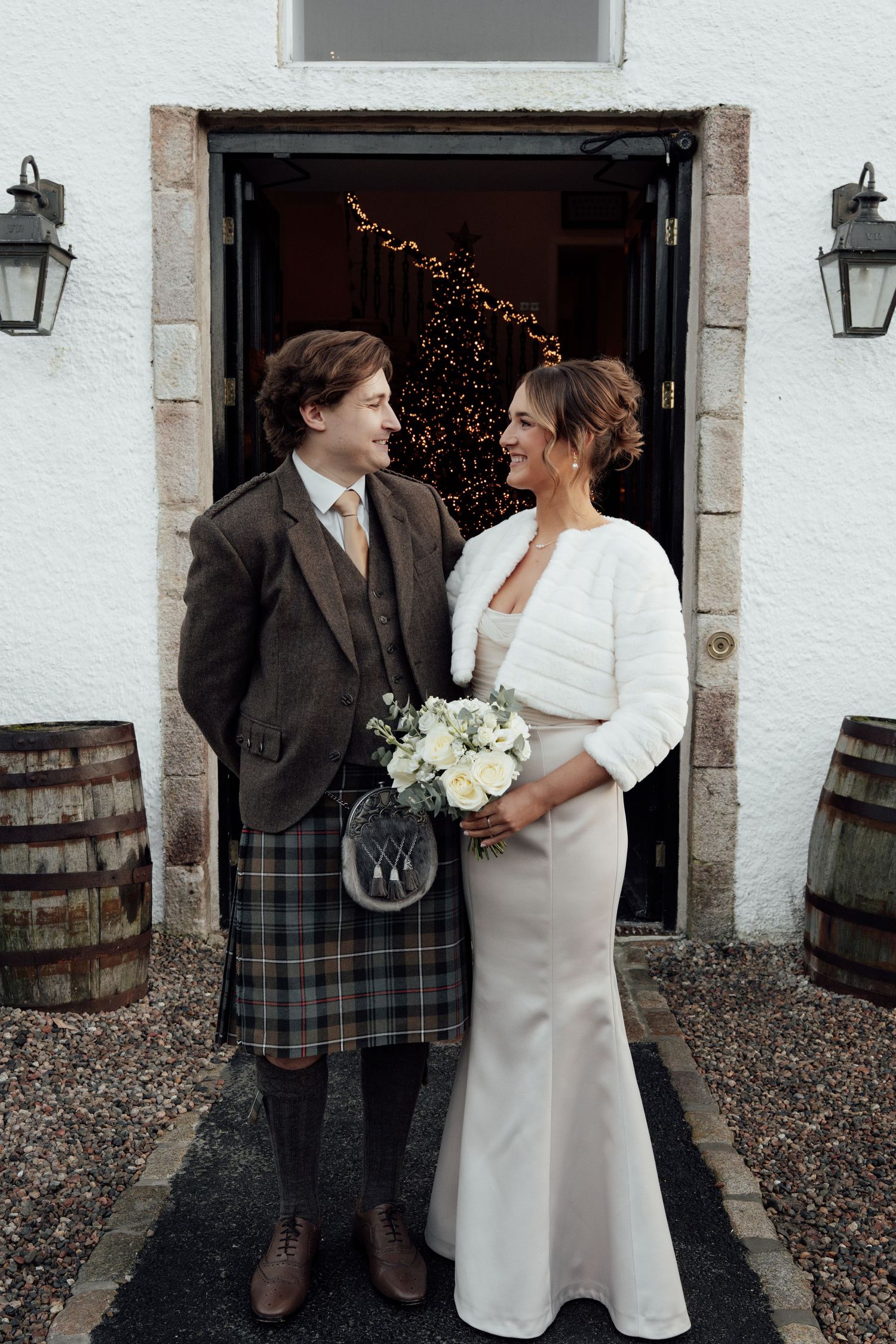 Wedding couple poses in front of white stone building with wooden barrels, groom in Scottish kilt.