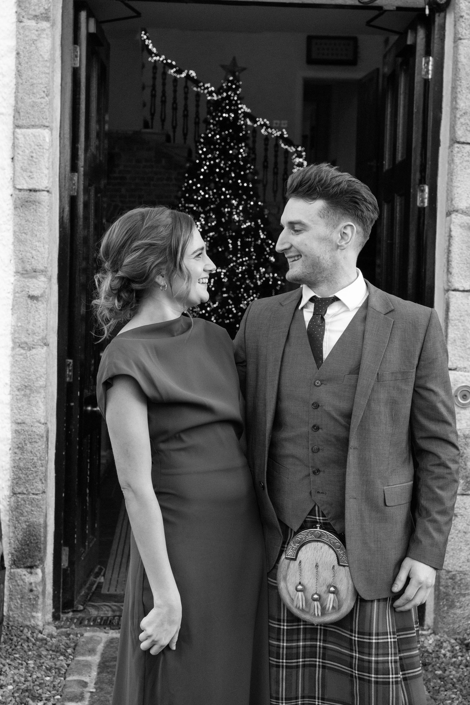 Black and white photo of a couple in formal attire standing by a doorway with twinkling lights.