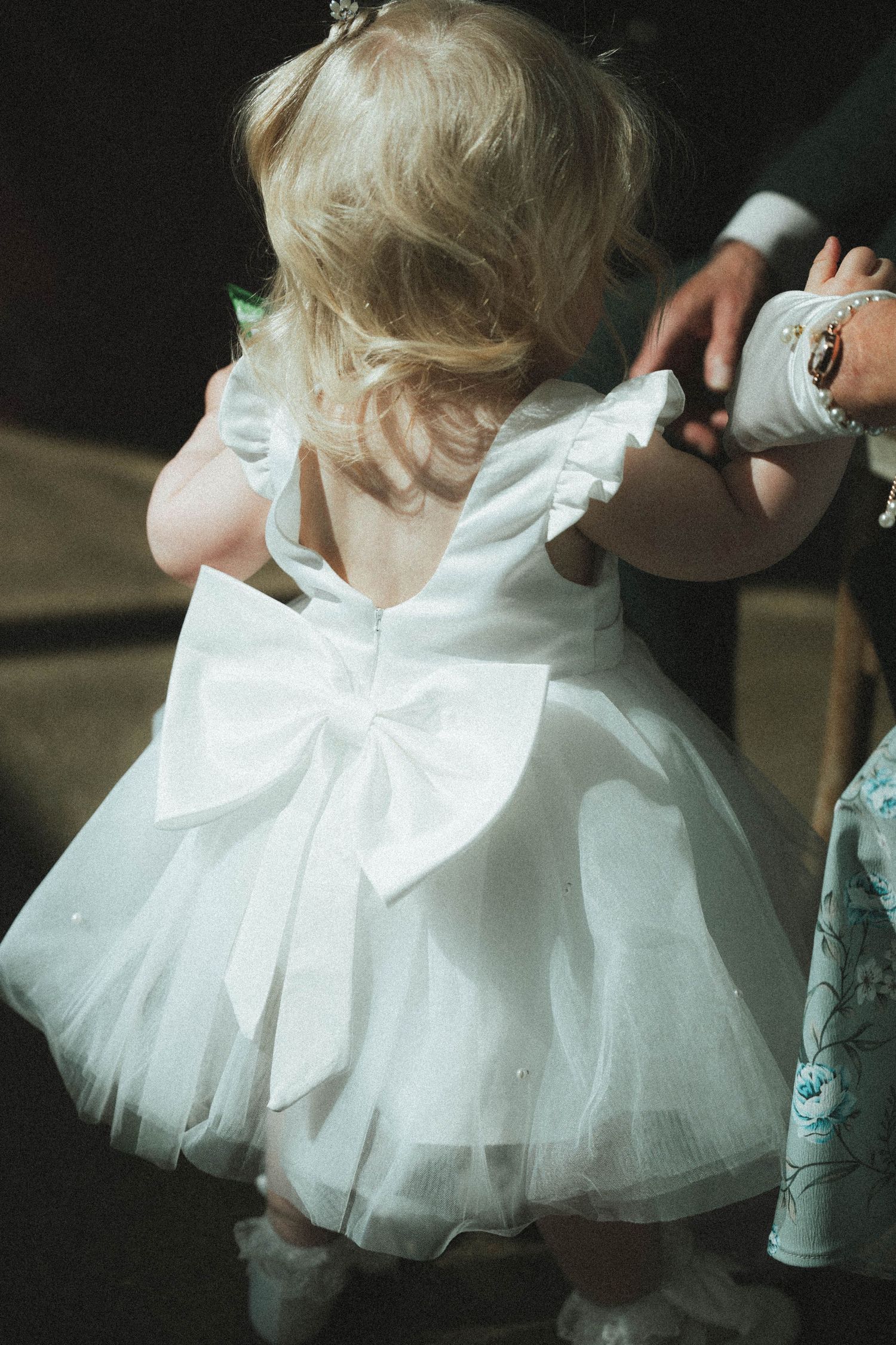 Adorable toddler flower girl in white tutu dress with large bow at back during wedding.