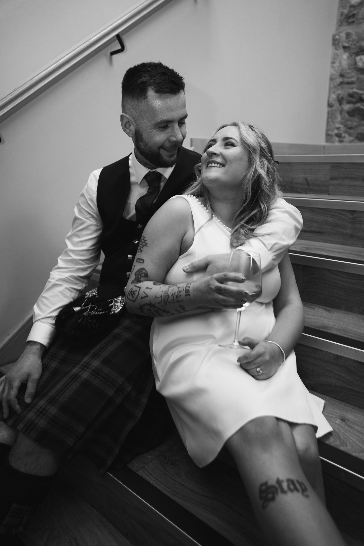 Black and white photo of tattooed bride and groom in kilt sitting on stairs, sharing a romantic moment.