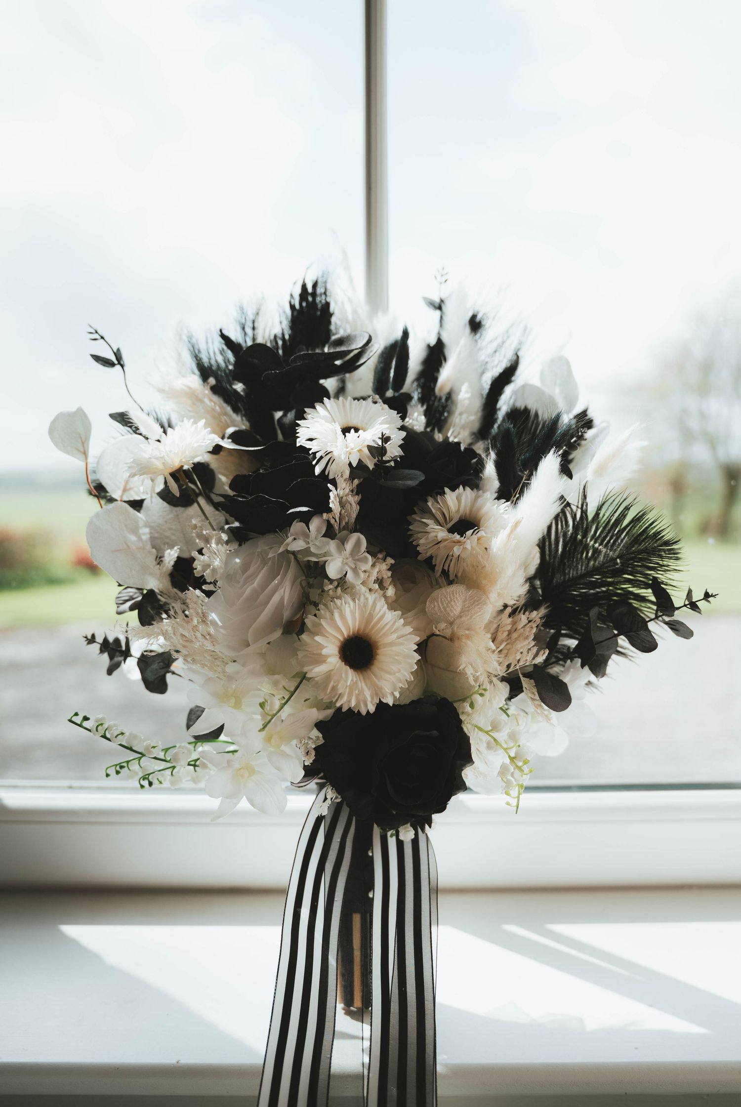 Elegant black and white wedding bouquet with gerberas, roses, and feathers tied with striped ribbon on windowsill.