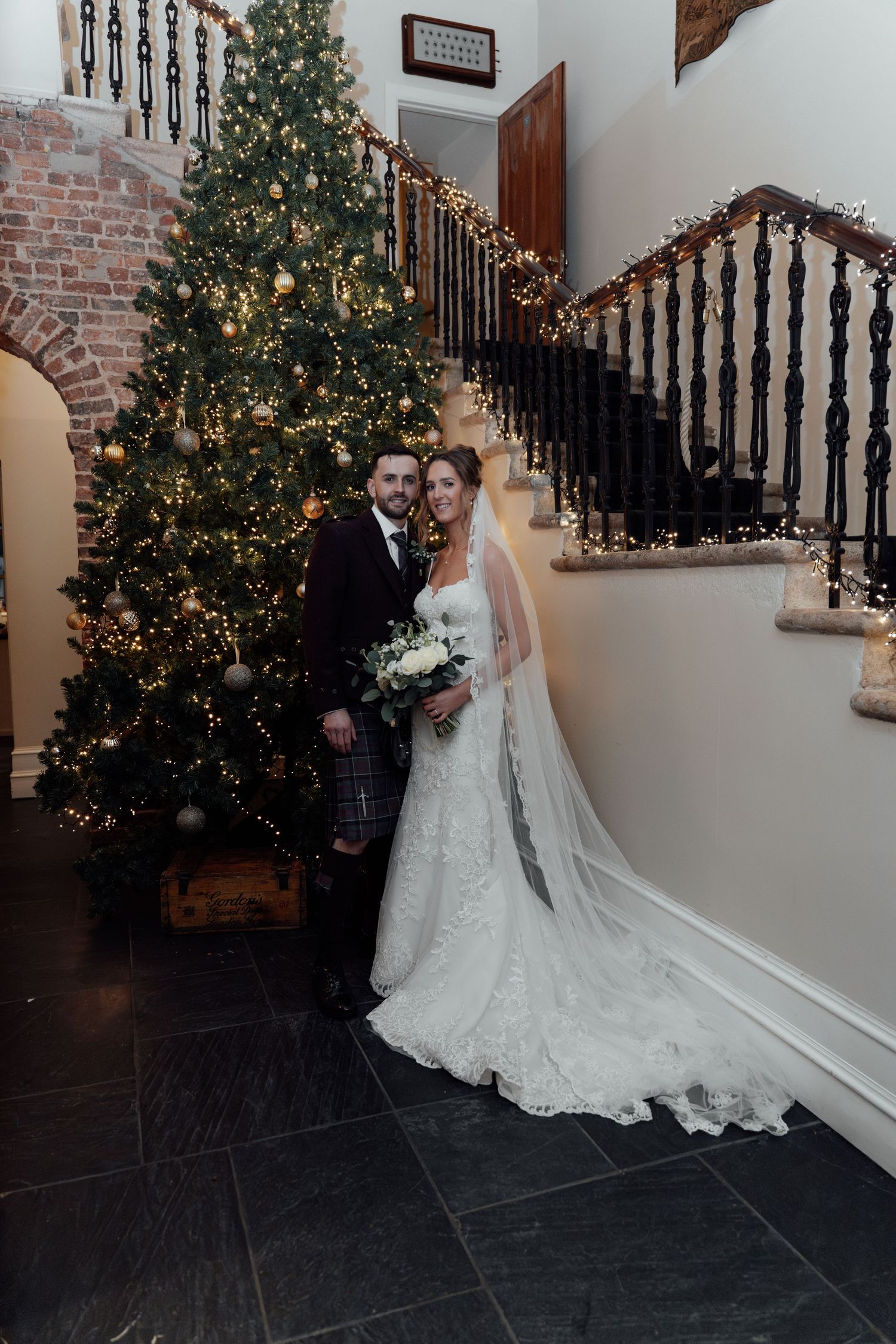 Full-length wedding portrait by Christmas tree with bride's train extending across slate floor.